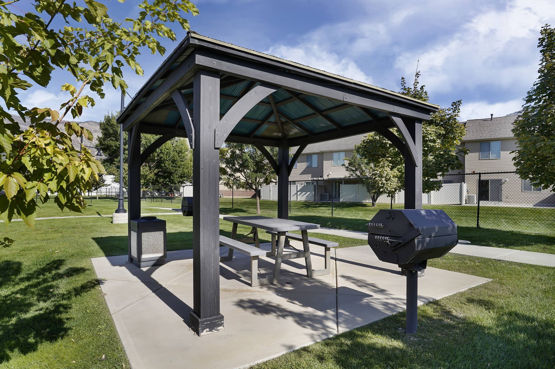 Picnic shelter with table, benches, grill, and trash can on concrete pad in a grassy area.