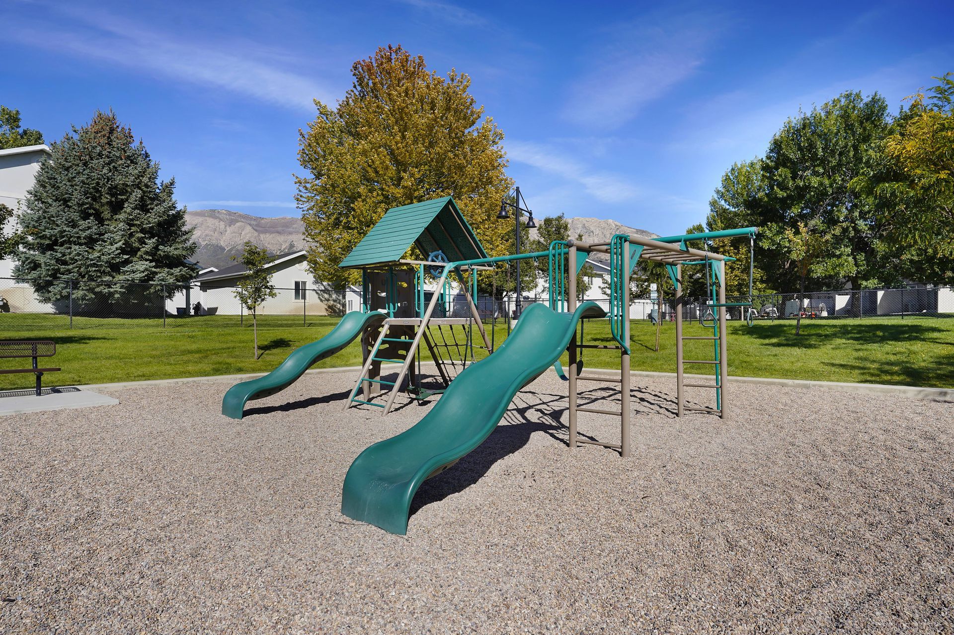 Playground with green slides, brown structure, wood chips, and trees under a blue sky.
