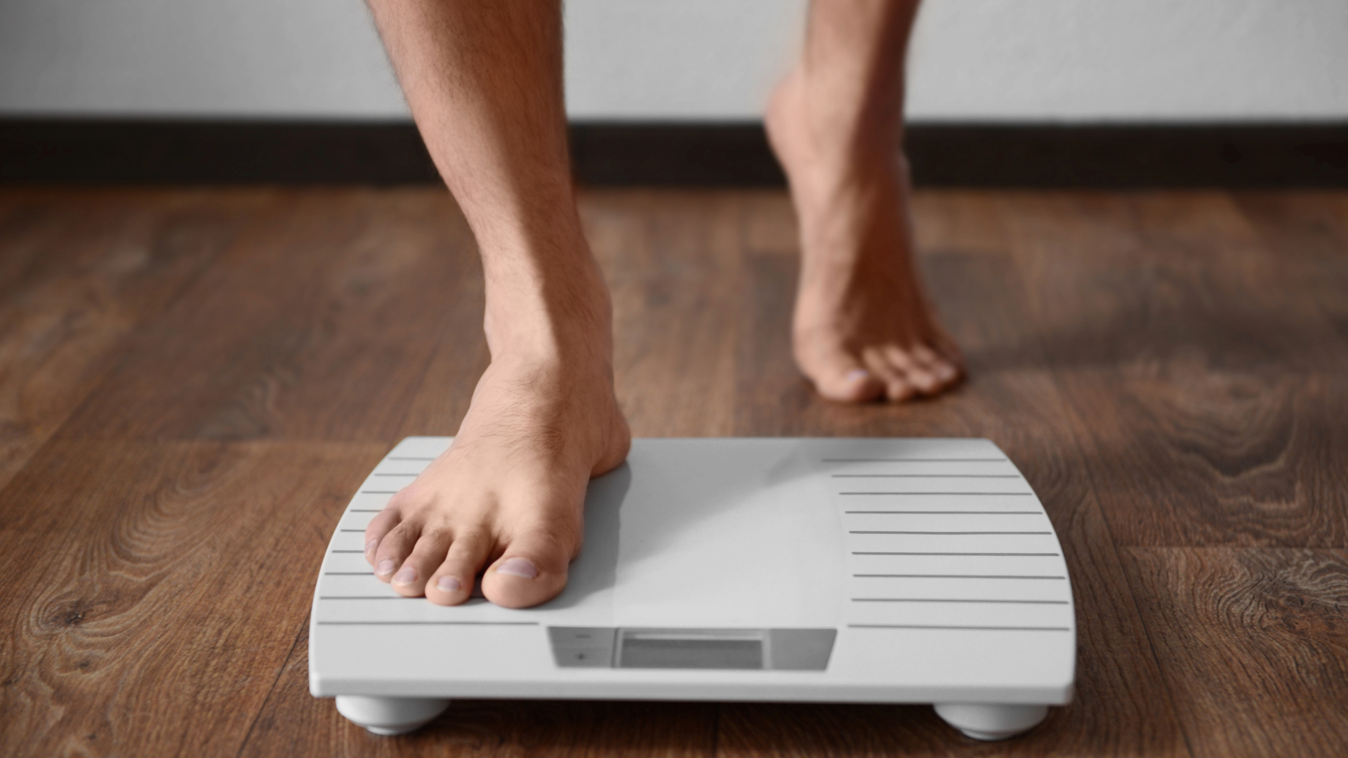 Person stepping on a white digital bathroom scale on a wooden floor.