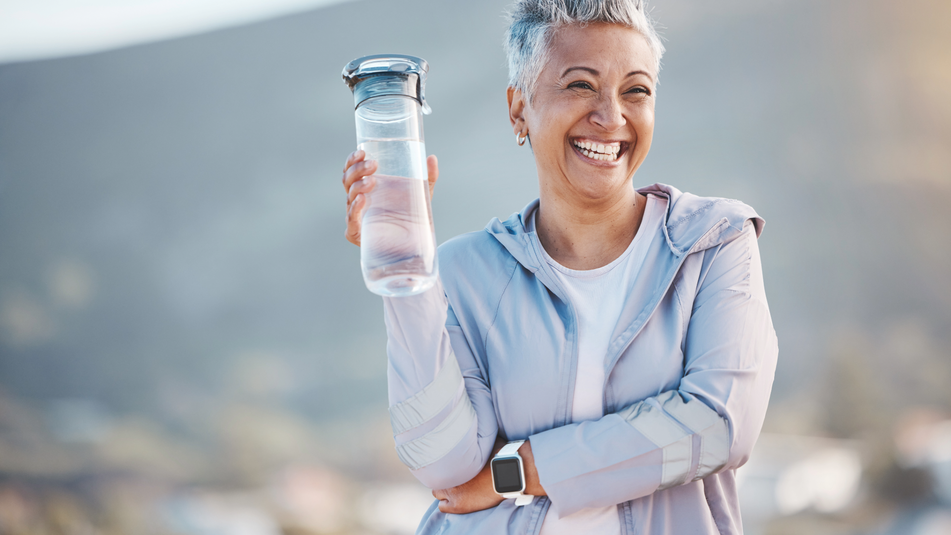 Smiling person in a light jacket holding a water bottle outdoors on a sunny day