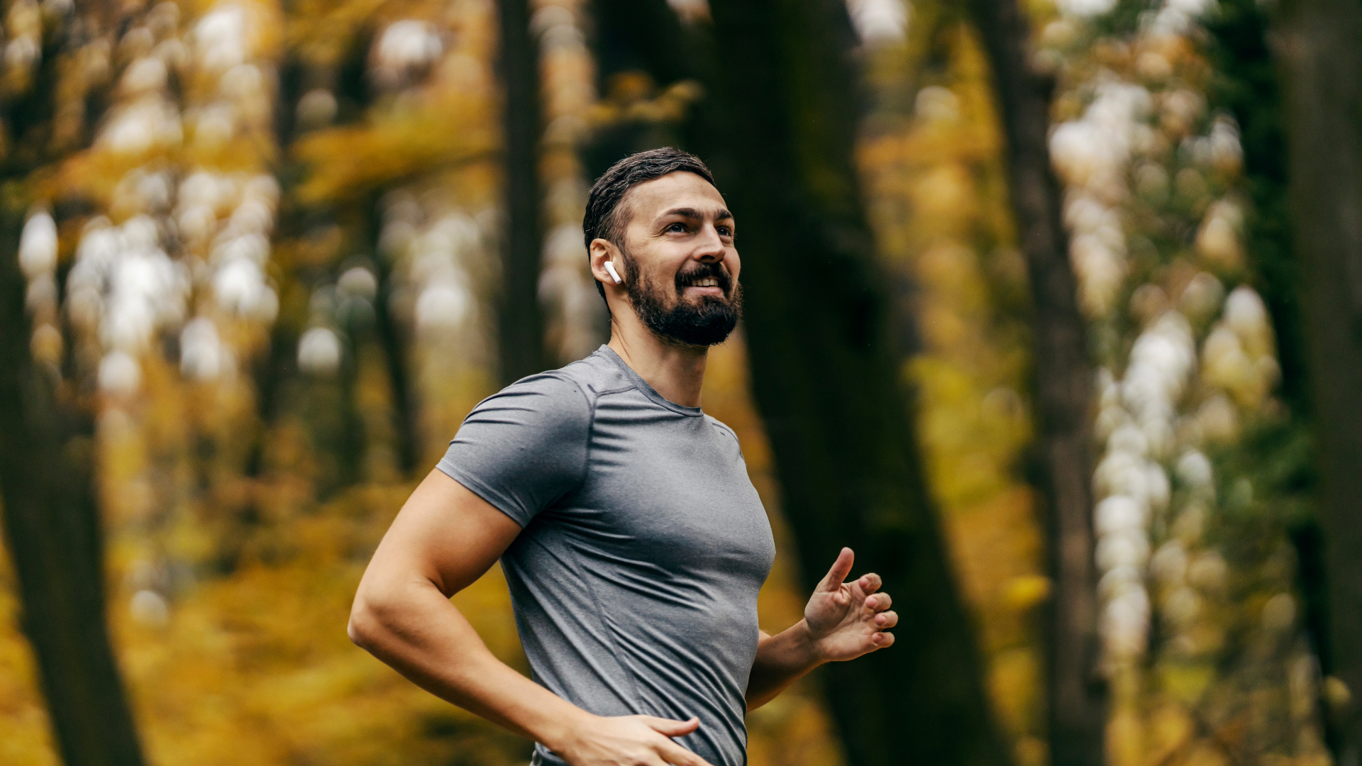 Man jogging in a forest trail, wearing a gray shirt, with autumn trees blurred in the background