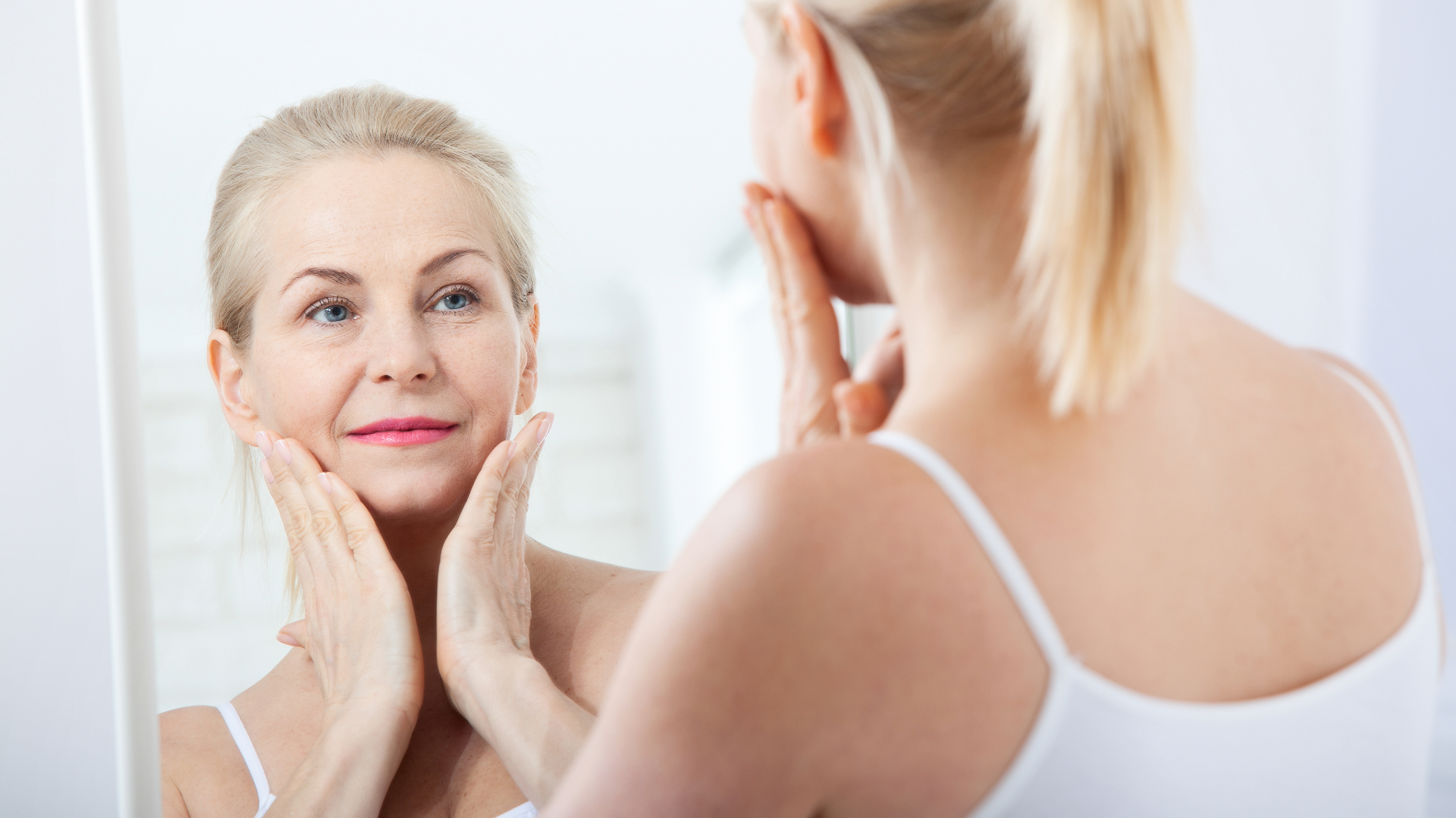 Woman examining her face in a mirror, gently touching her cheeks in a bright bathroom.
