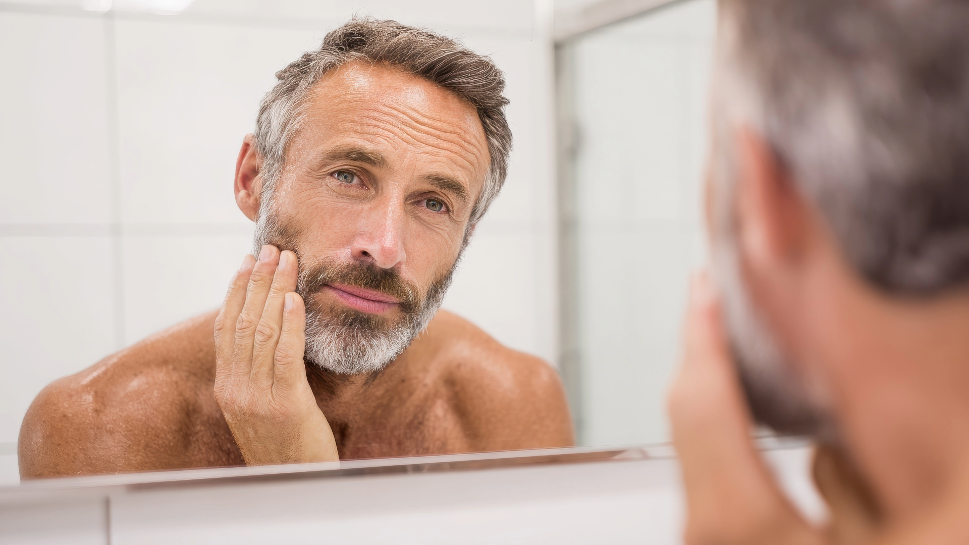 Man examining his face in a bathroom mirror while touching his cheek