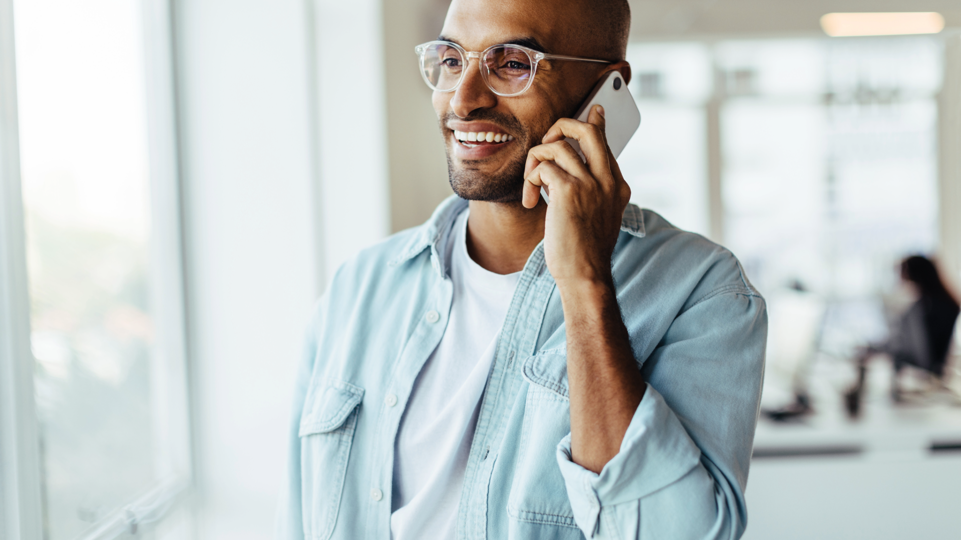 Man smiling while talking on a phone in a bright office, wearing glasses and a light blue shirt