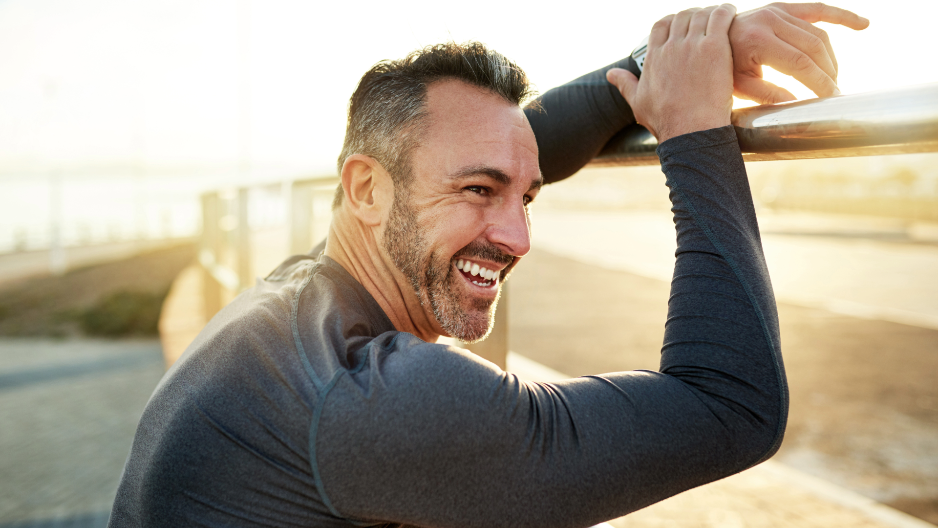 Smiling man in dark athletic shirt leaning on a railing outdoors in warm sunlight