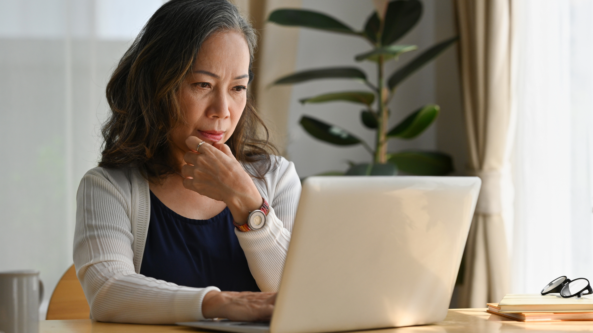 Woman seated at a laptop, resting her chin on her hand in a bright home office
