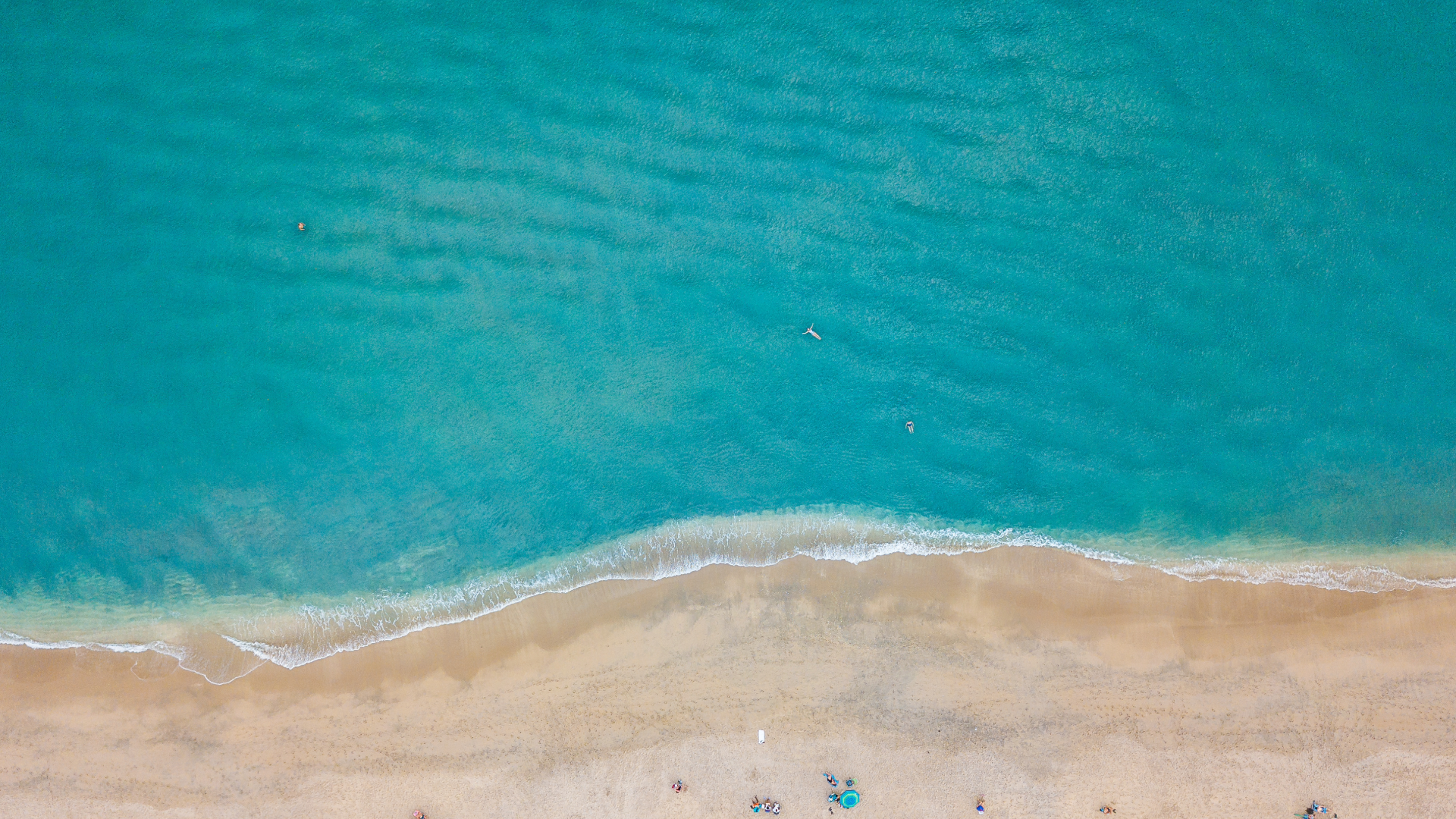 Aerial view of turquoise ocean meeting a sandy beach with small people near the shoreline