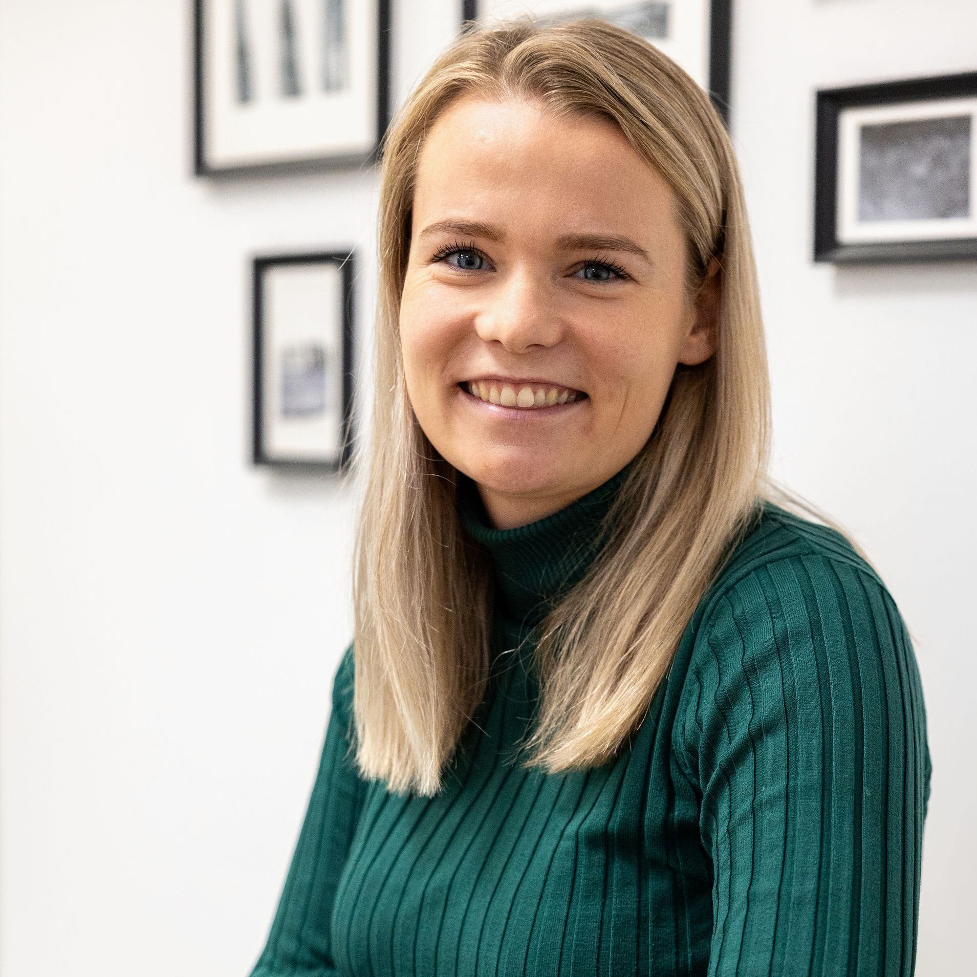 Een blonde vrouw, gekleed in een groene coltrui, glimlacht voor een ingelijst kunstwerk aan een witte muur.