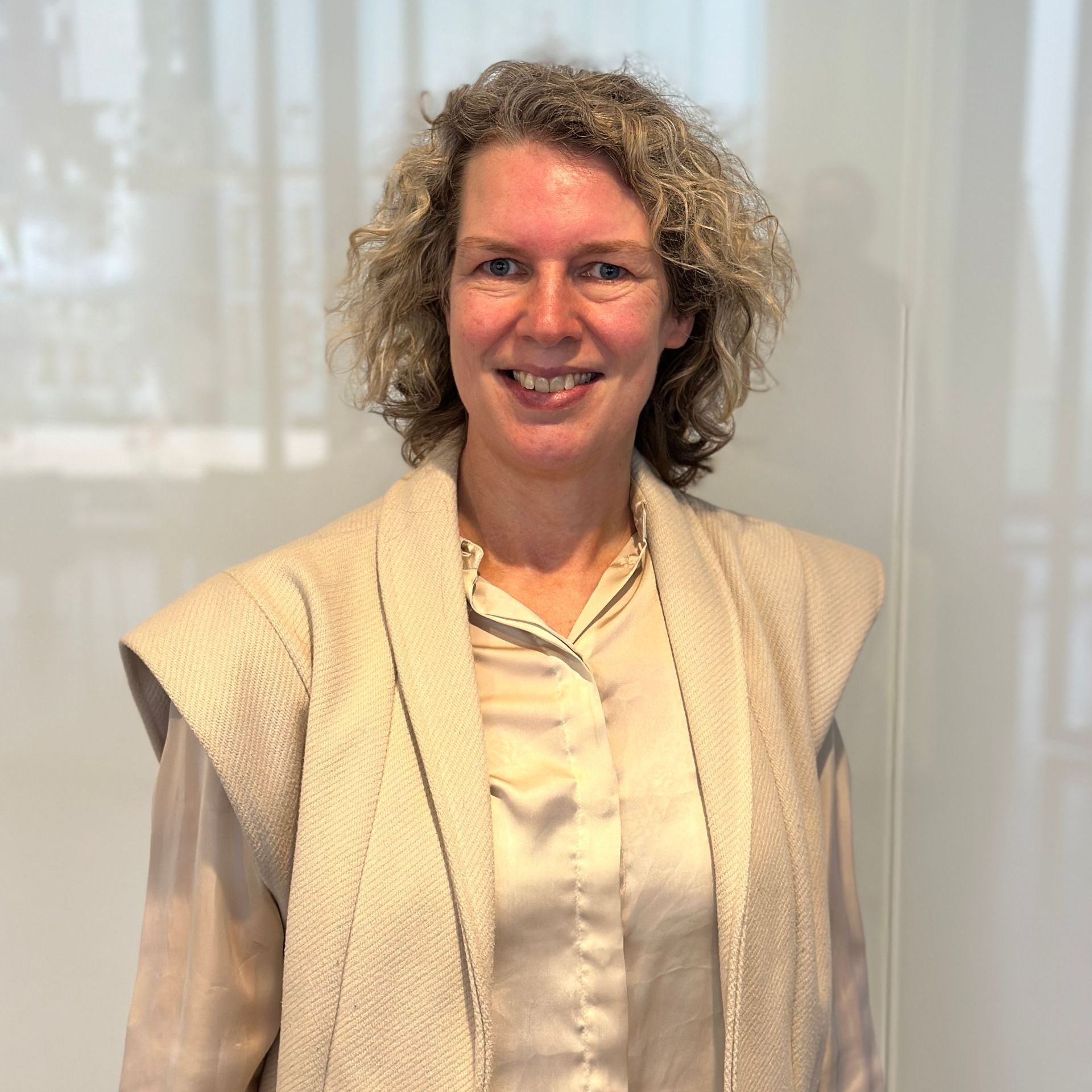 Een vrouw met krullend haar, gekleed in een beige vest en blouse, staat binnen en lacht.