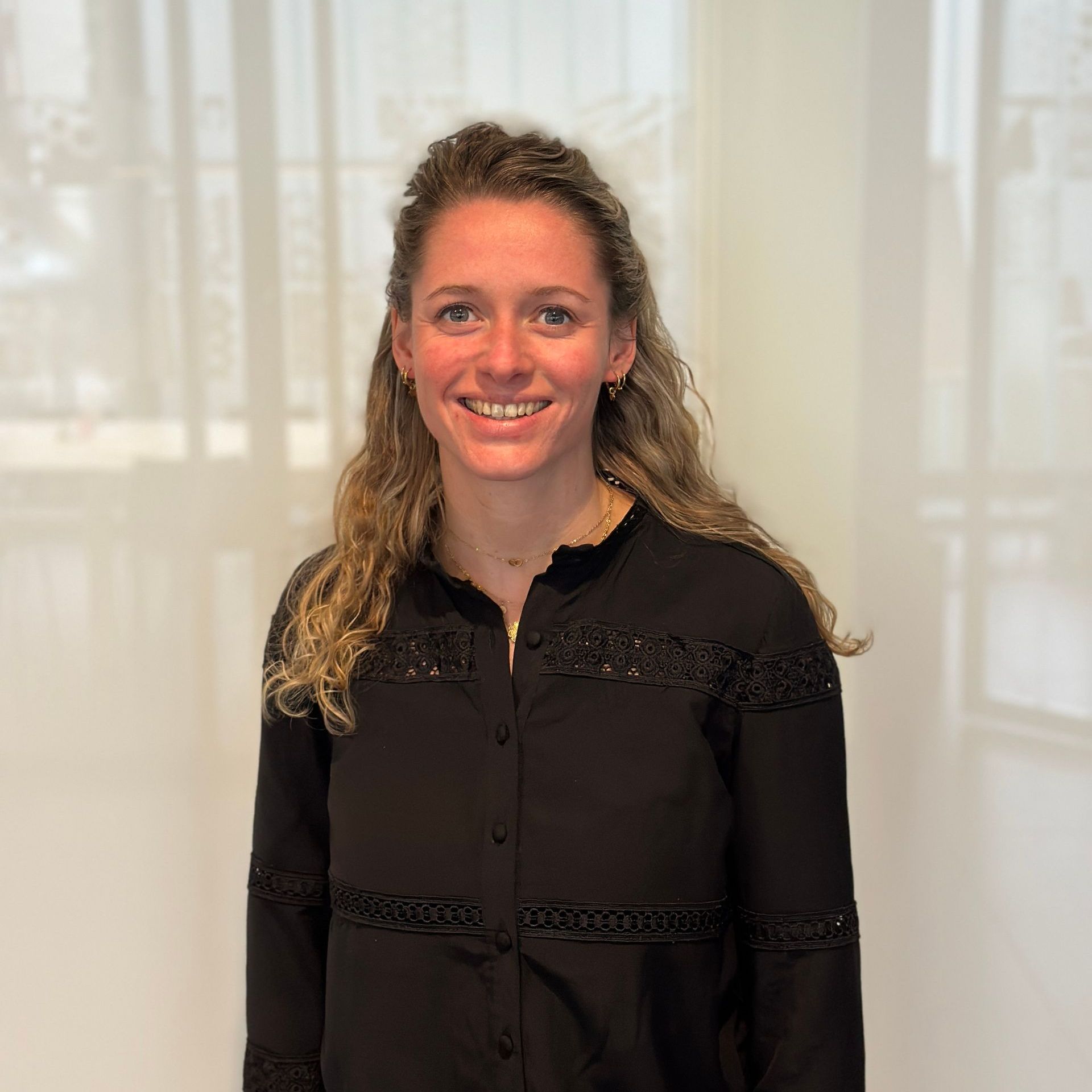 Een lachende vrouw, gekleed in een zwart shirt, staat binnen bij een raam.