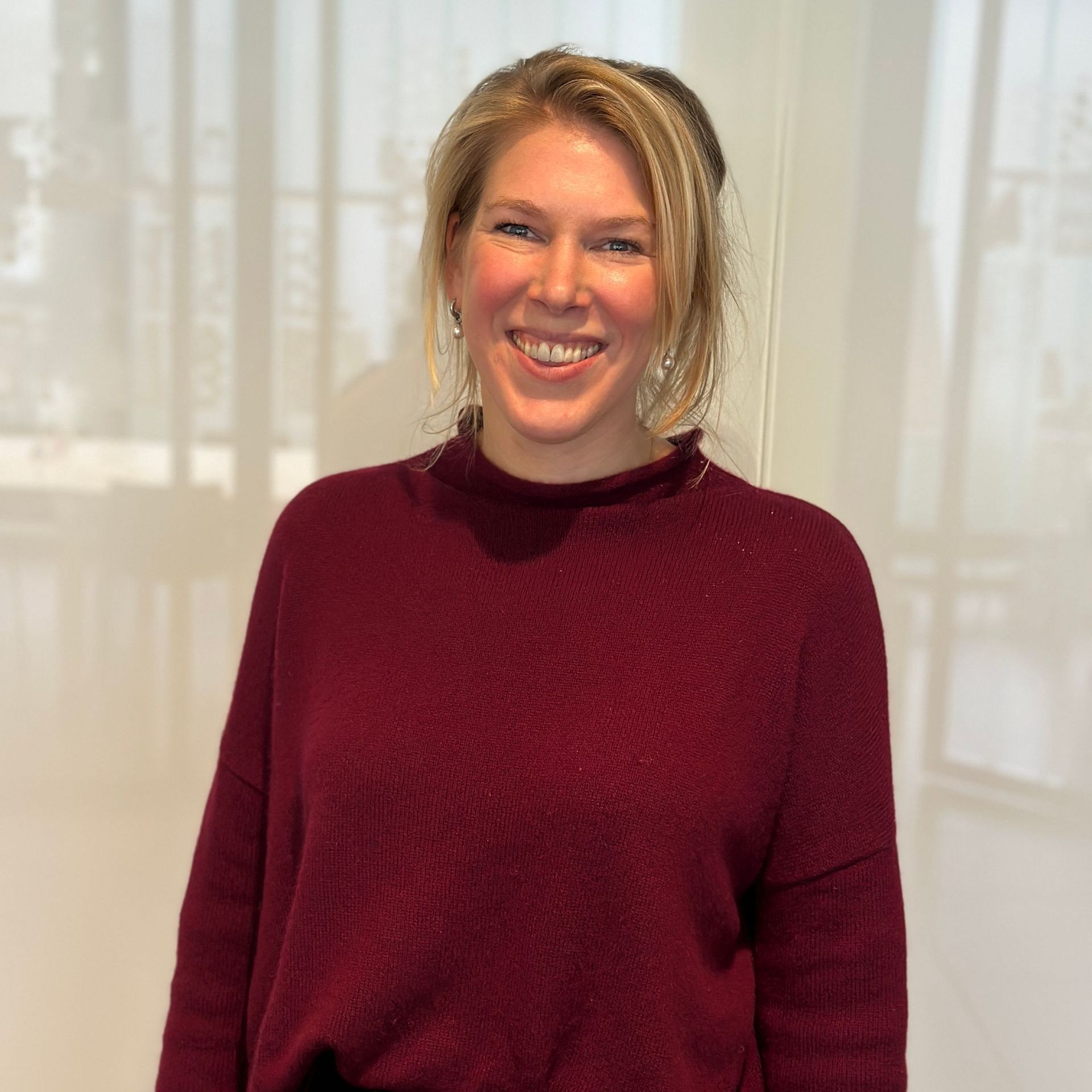 Vrouw met blond haar, gekleed in een bordeauxrode trui, lacht. Ze staat bij een raam.