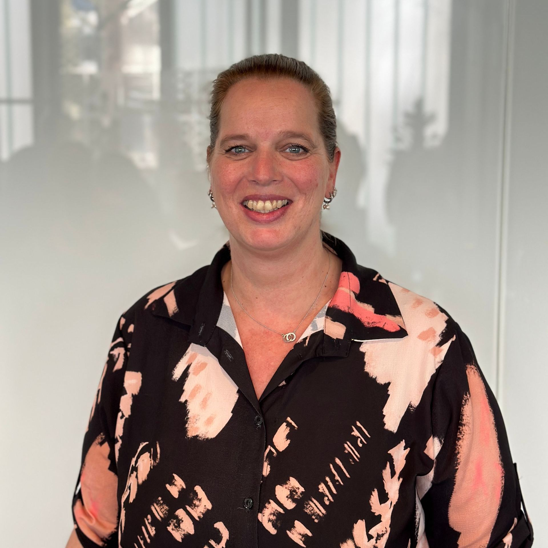 Een vrouw met een lichte huid, gekleed in een zwart shirt met een abstract roze en wit patroon, lacht in een lichte kamer.