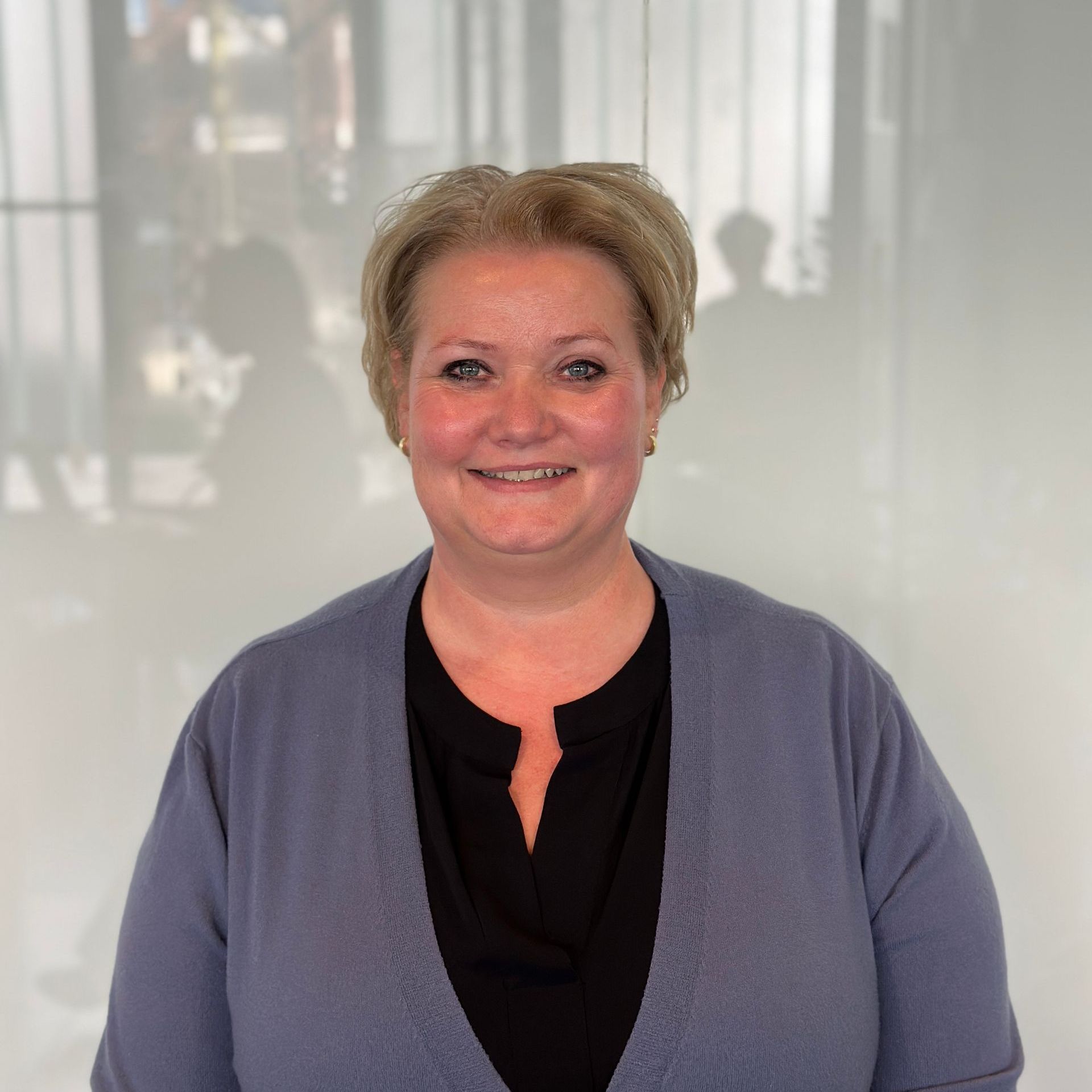 Een lachende vrouw, gekleed in een blauw vest over een zwart shirt, in een moderne kantooromgeving.
