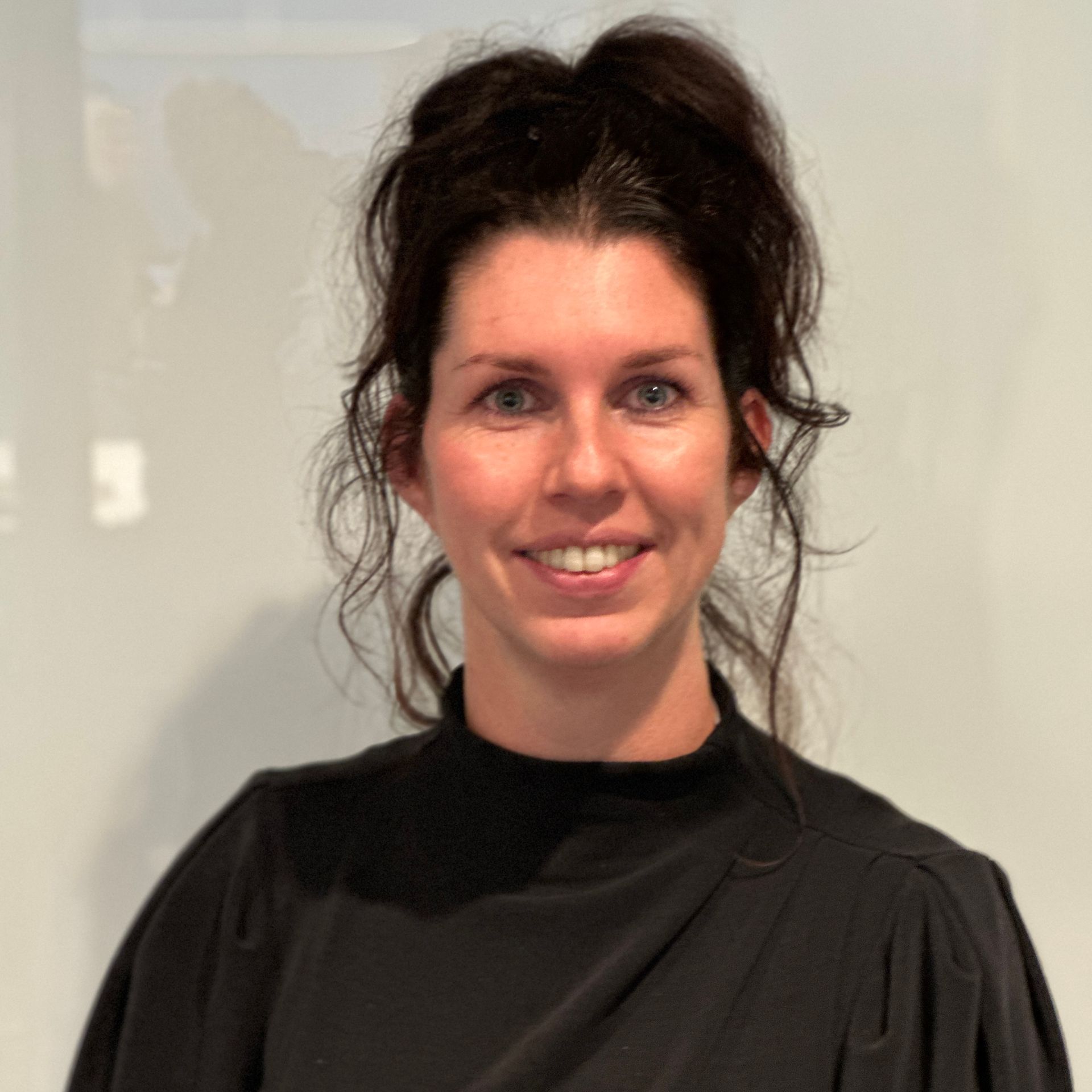Vrouw met donker haar, gekleed in een zwarte top, lacht tegen een witte muur.