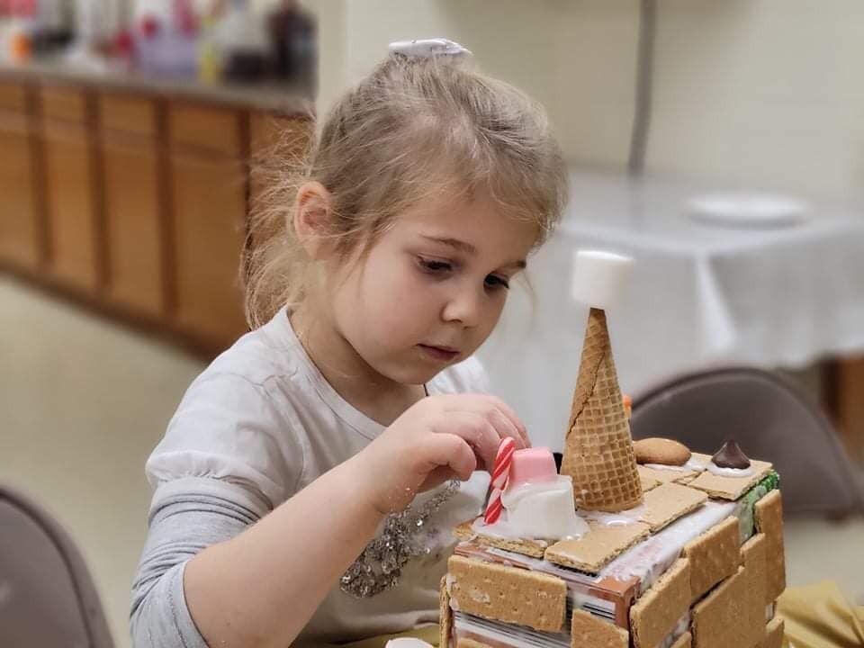 Child carefully decorating a small cake with frosting and a waffle cone, seated at a table indoors.