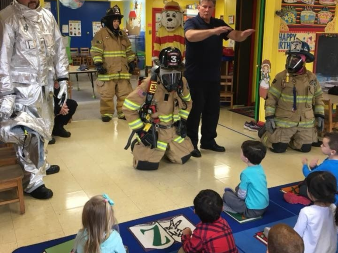 Firefighters in gear demonstrate for children sitting on a classroom carpet.