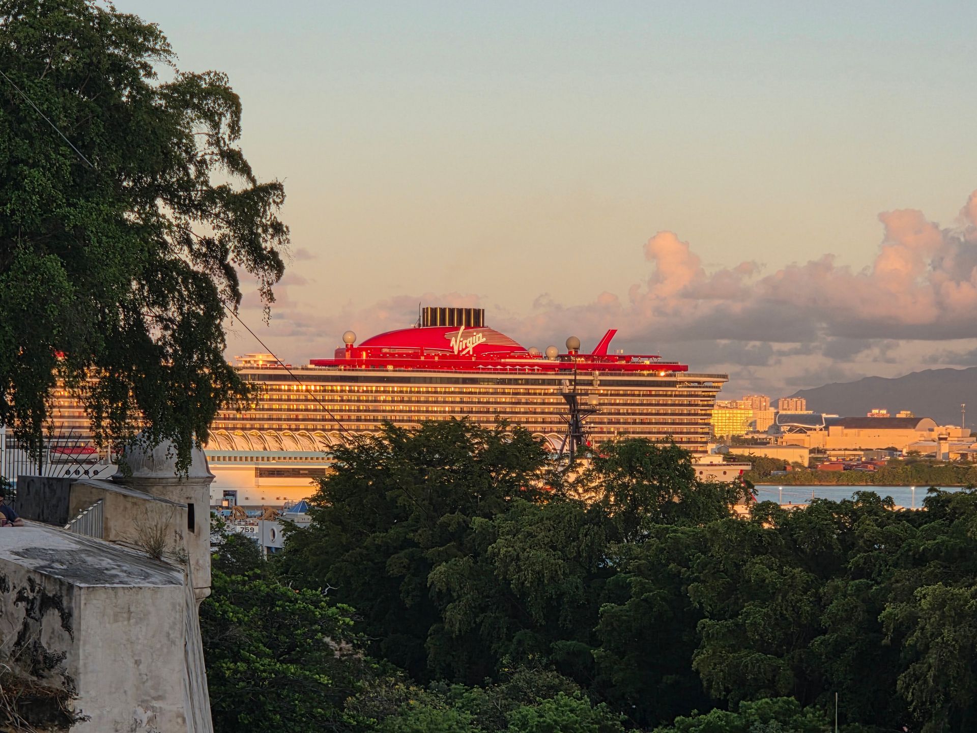 Cruise ship with bright red lights at sunset, seen beyond trees and hills.