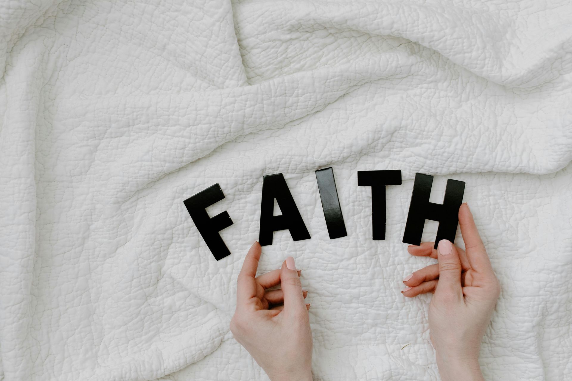 Hands arranging black letters spelling “FAITH” on a white textured blanket.