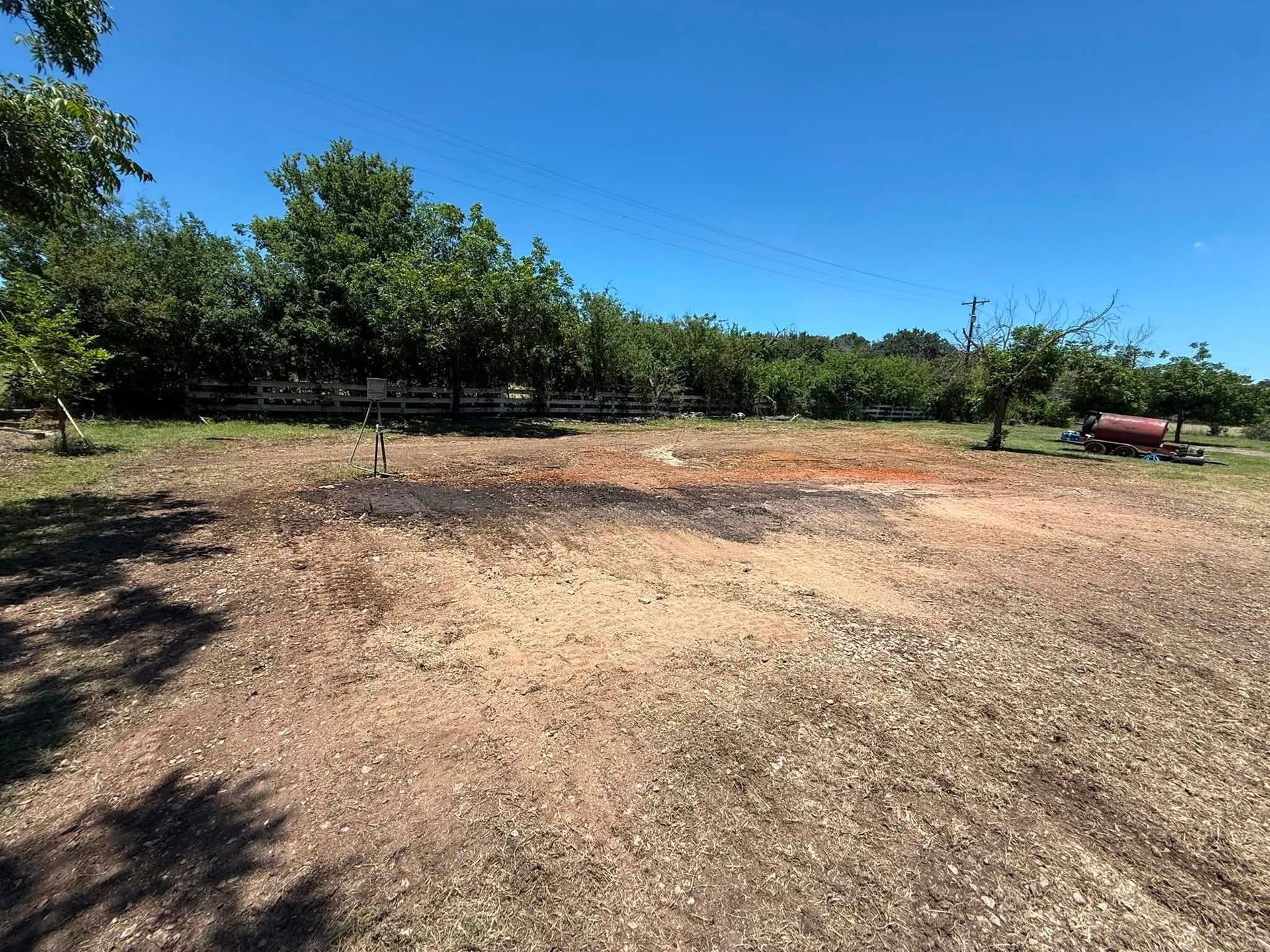 Open dirt field with trees under a clear blue sky, and a red tank at the far right.