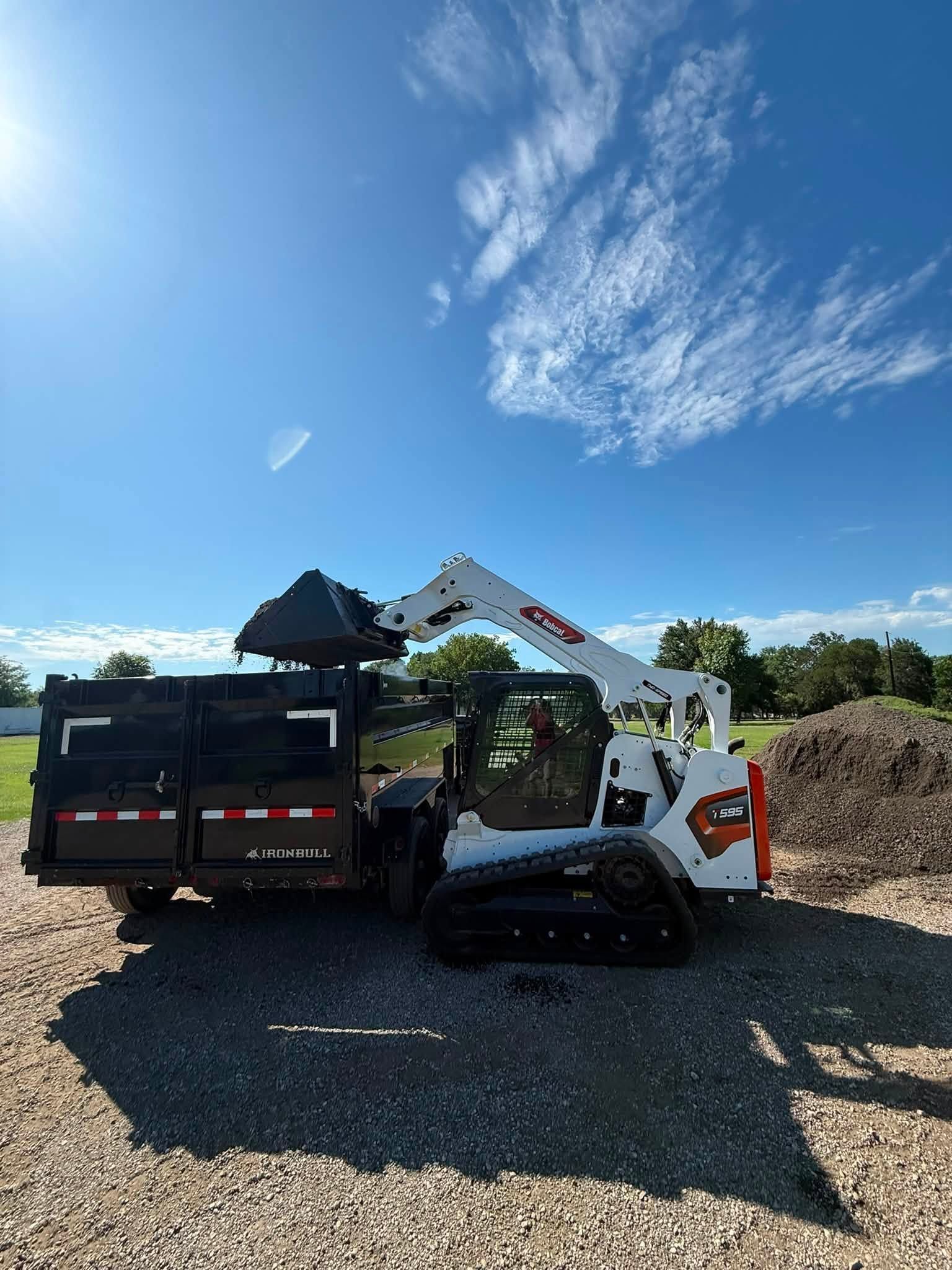 White skid steer loader beside a black dump truck on a gravel lot under a bright blue sky.