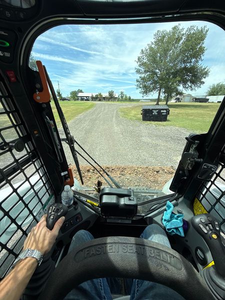 Inside a skid steer cab on a gravel path, looking toward a grassy yard with a tree and a black container.