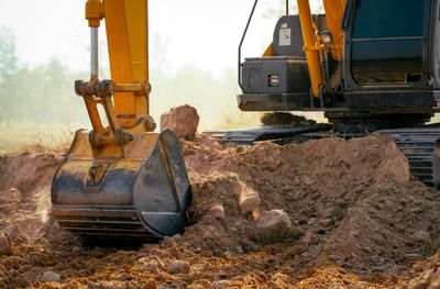Yellow excavator bucket digging dirt at a construction site, with soil piled beside the machine.