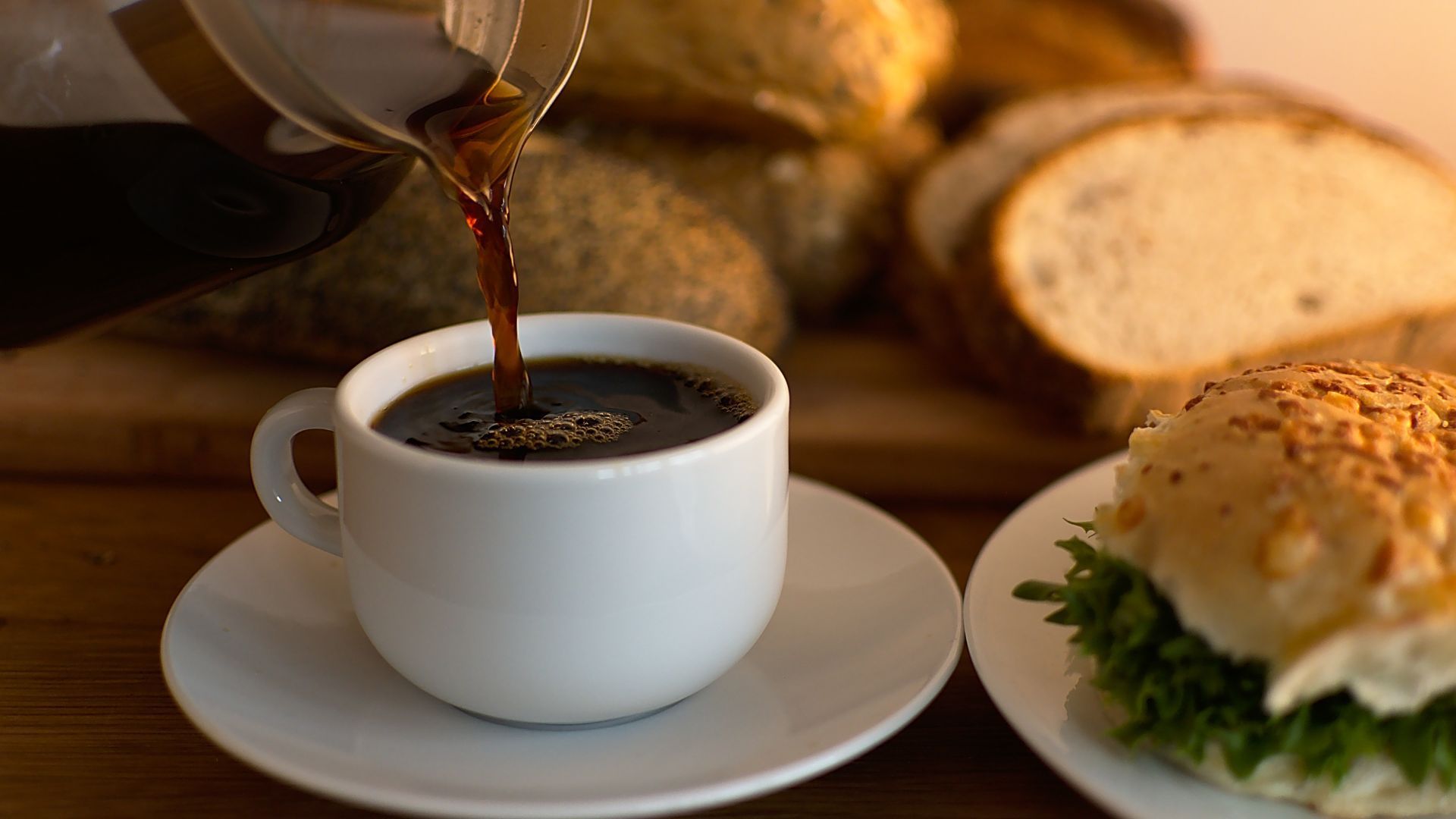 Kaffee wird in eine weiße Tasse eingeschenkt. Im Hintergrund sieht man frisches Brot und Backwaren.