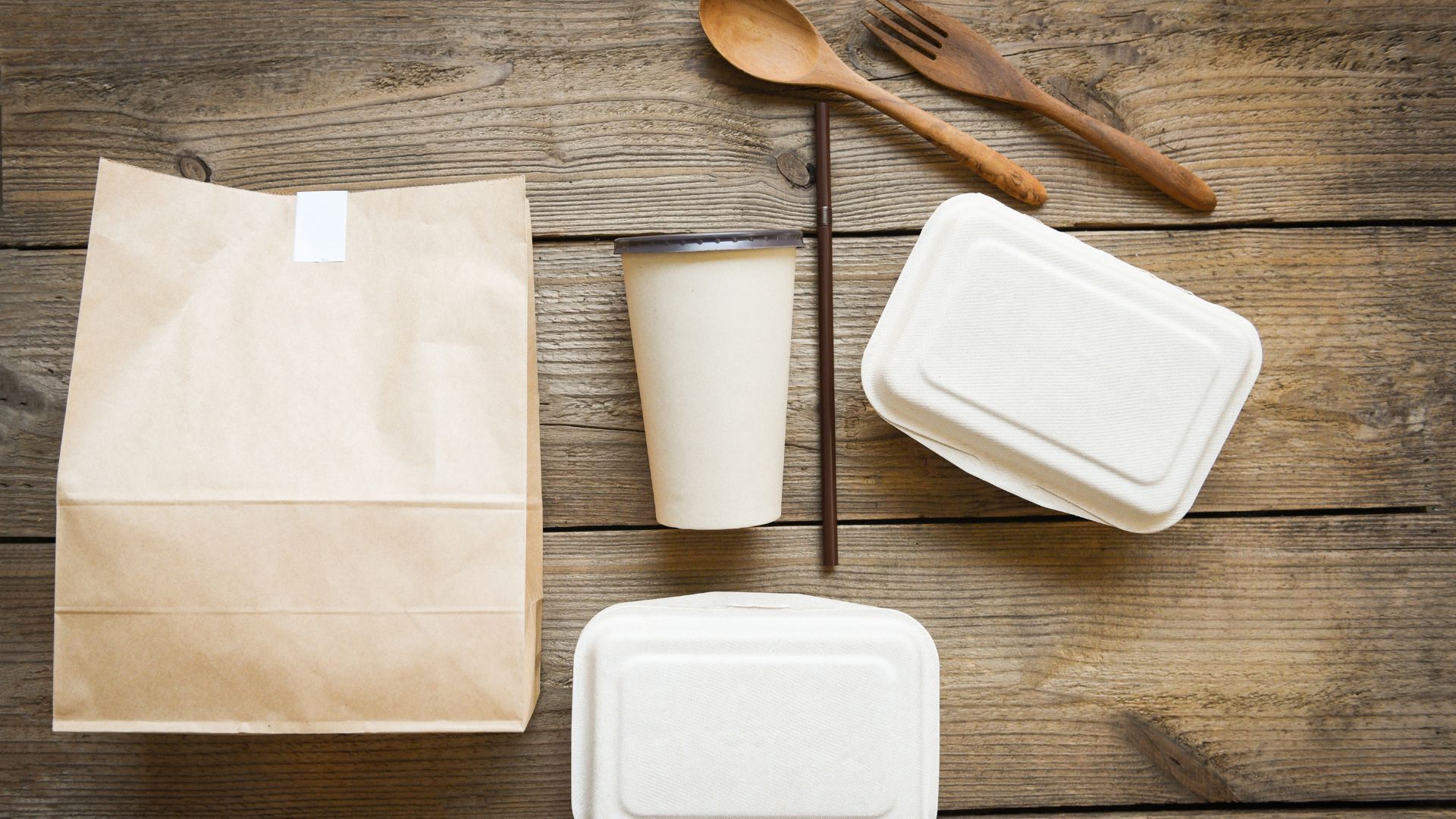 Auf einem Holztisch liegen Verpackungen, die Essen zum Mitnehmen enthalten. Daneben liegen eine braune Tüte, Pappbecher etc