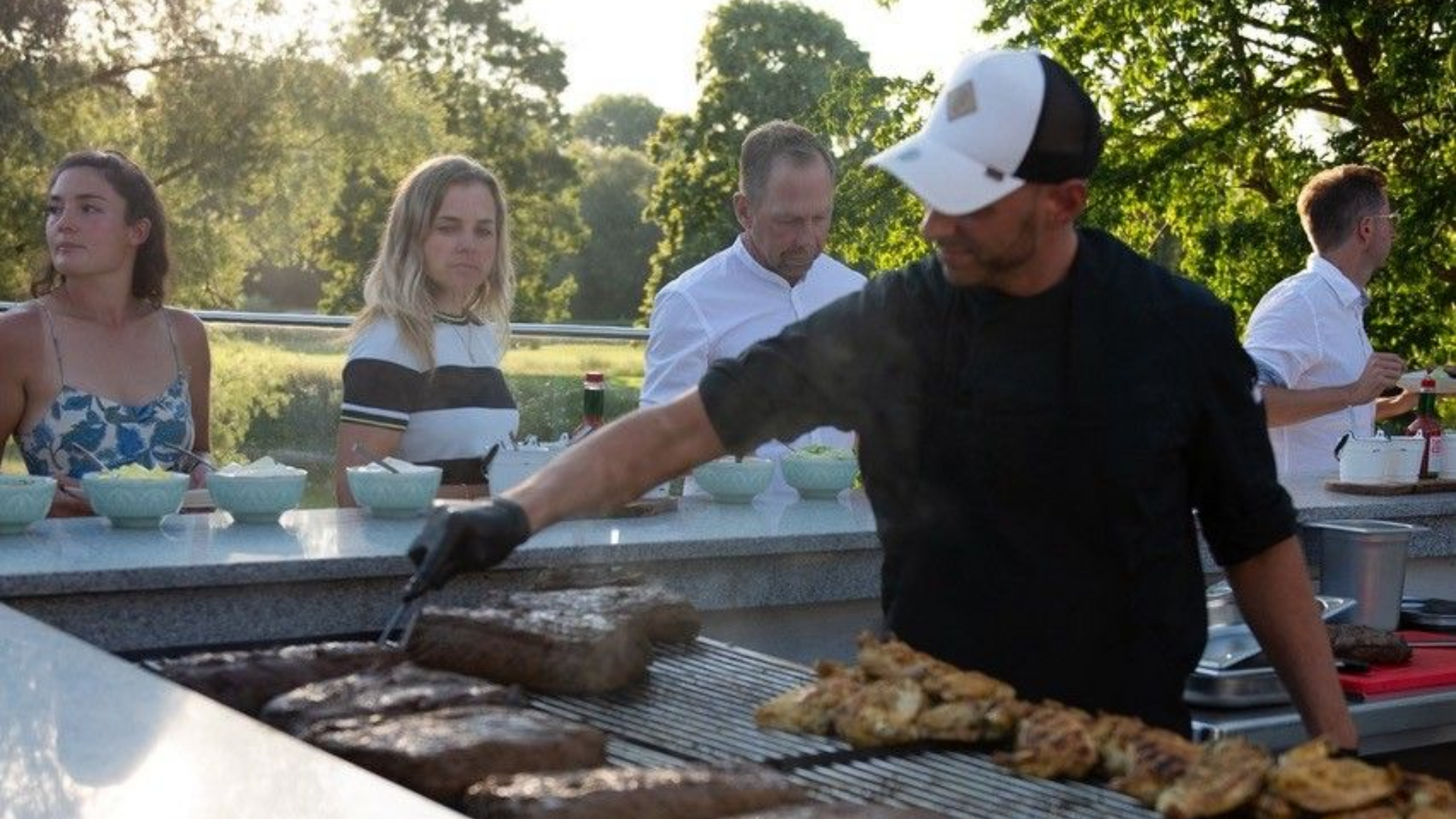 Simon Bartelt steht am Grill auf der Terrasse des Restaurants Platzhirsch und wendet Fleisch, während Gäste sich ihr Menü zusammenstellen