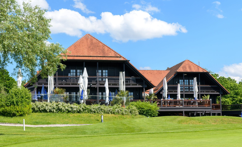Blick vom Golfplatz auf das Restaurant Platzhirsch in Olching mit Terrasse und Sonnenschirmen.