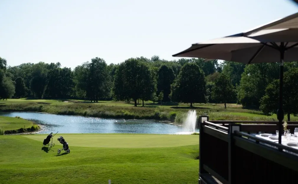 Blick von der Terrasse des Restaurants Platzhirsch auf den Golfplatz Olching mit See und Bäumen.