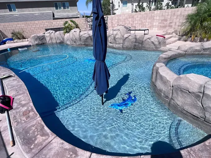 Swimming pool with blue water, surrounded by stone features and a patio. An umbrella casts a shadow.