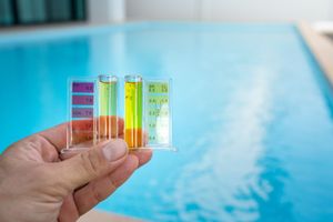 Hand holding pool water test kit with tubes of colored liquid against a blue pool.