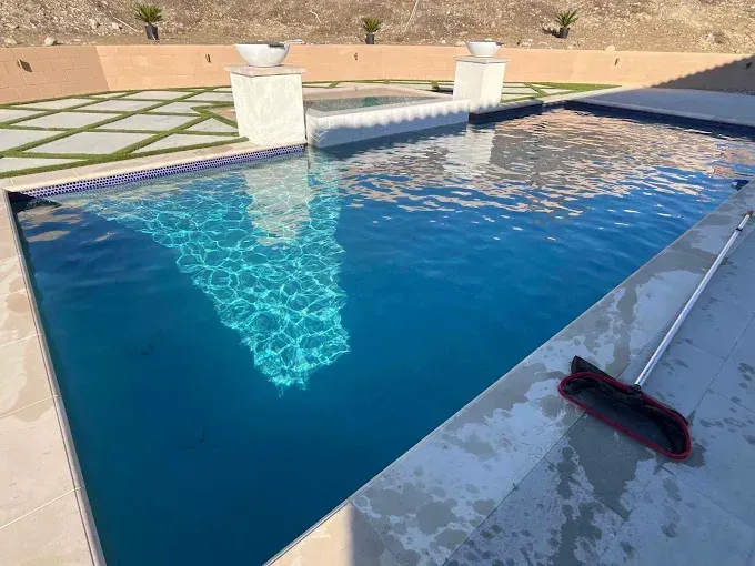 Pool with blue water and sunlight reflections, cleaner brush rests on the edge.