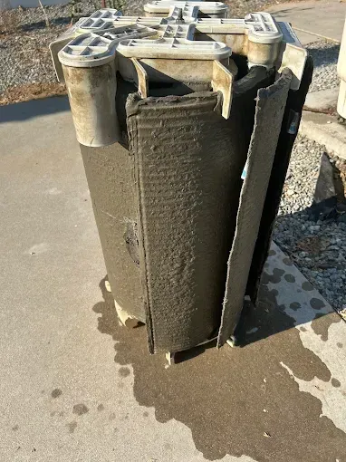 A cylindrical, dirty, gray filter on concrete. Parts are dislodged, and water is visible.