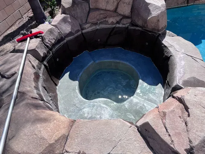 A circular hot tub built into rocky landscape next to a pool. A pool brush leans nearby.