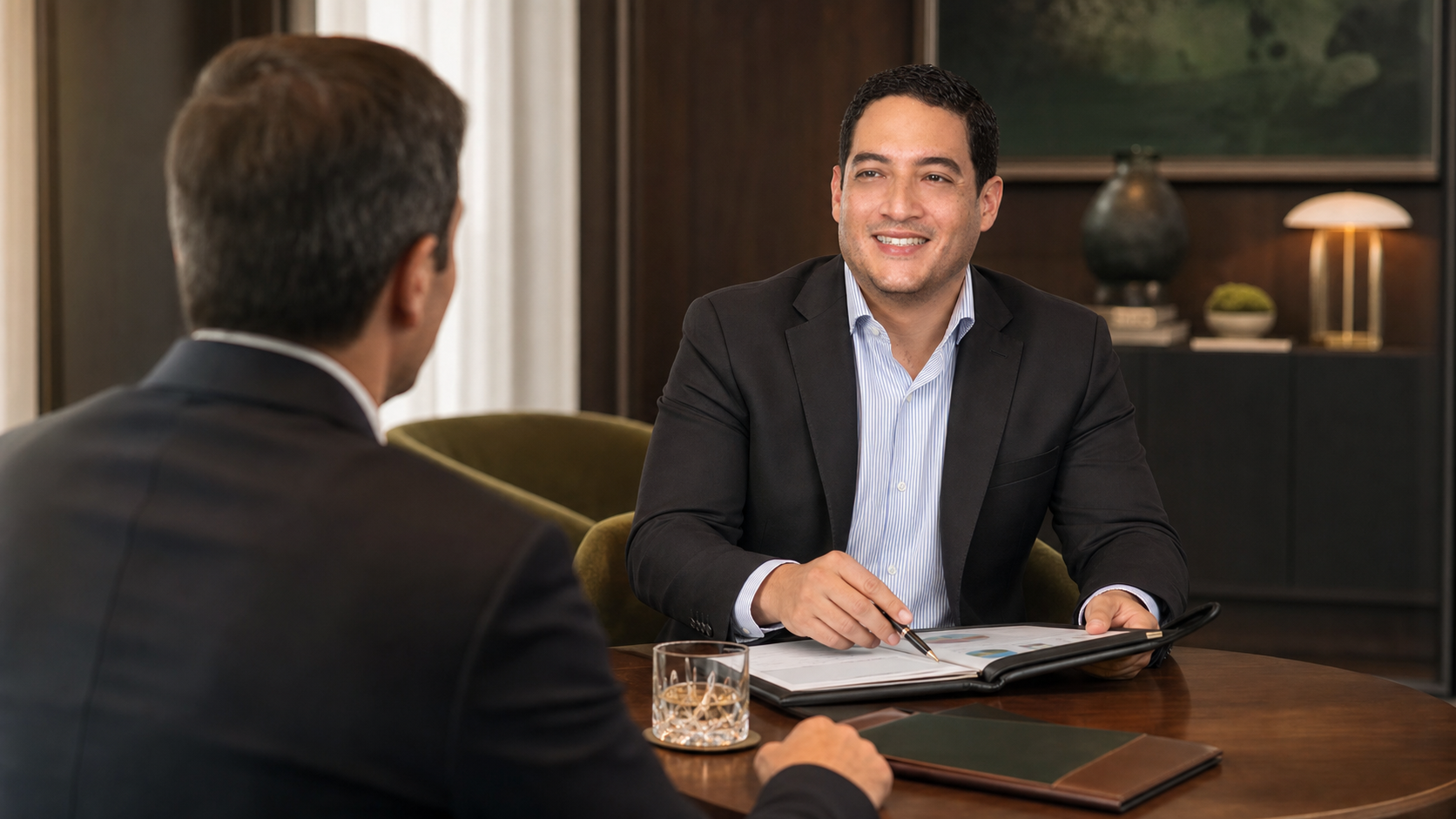Two men in business attire talking across a desk in a modern office, one smiling and holding a folder