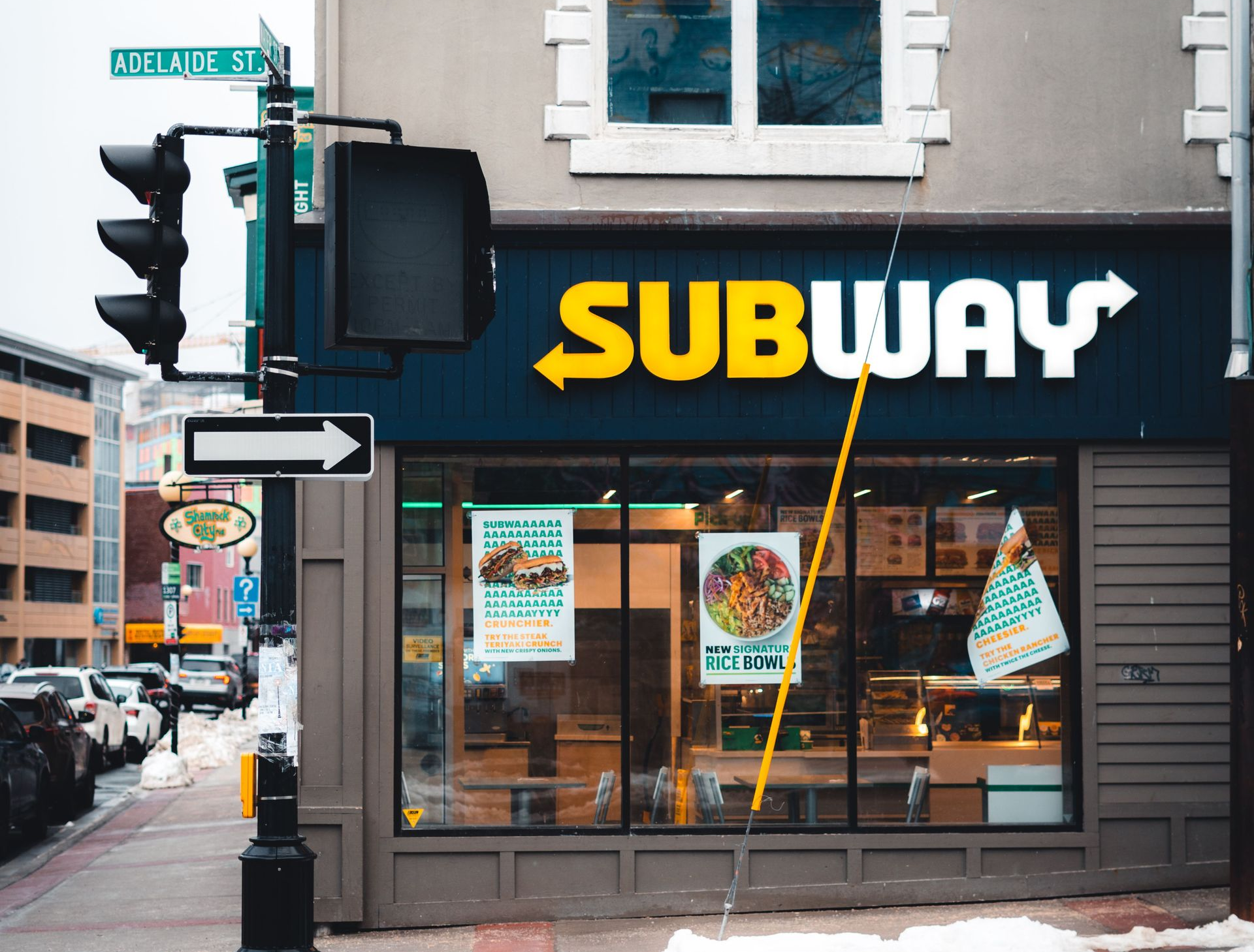 Subway restaurant storefront on a snowy city street with bold yellow sign and traffic lights