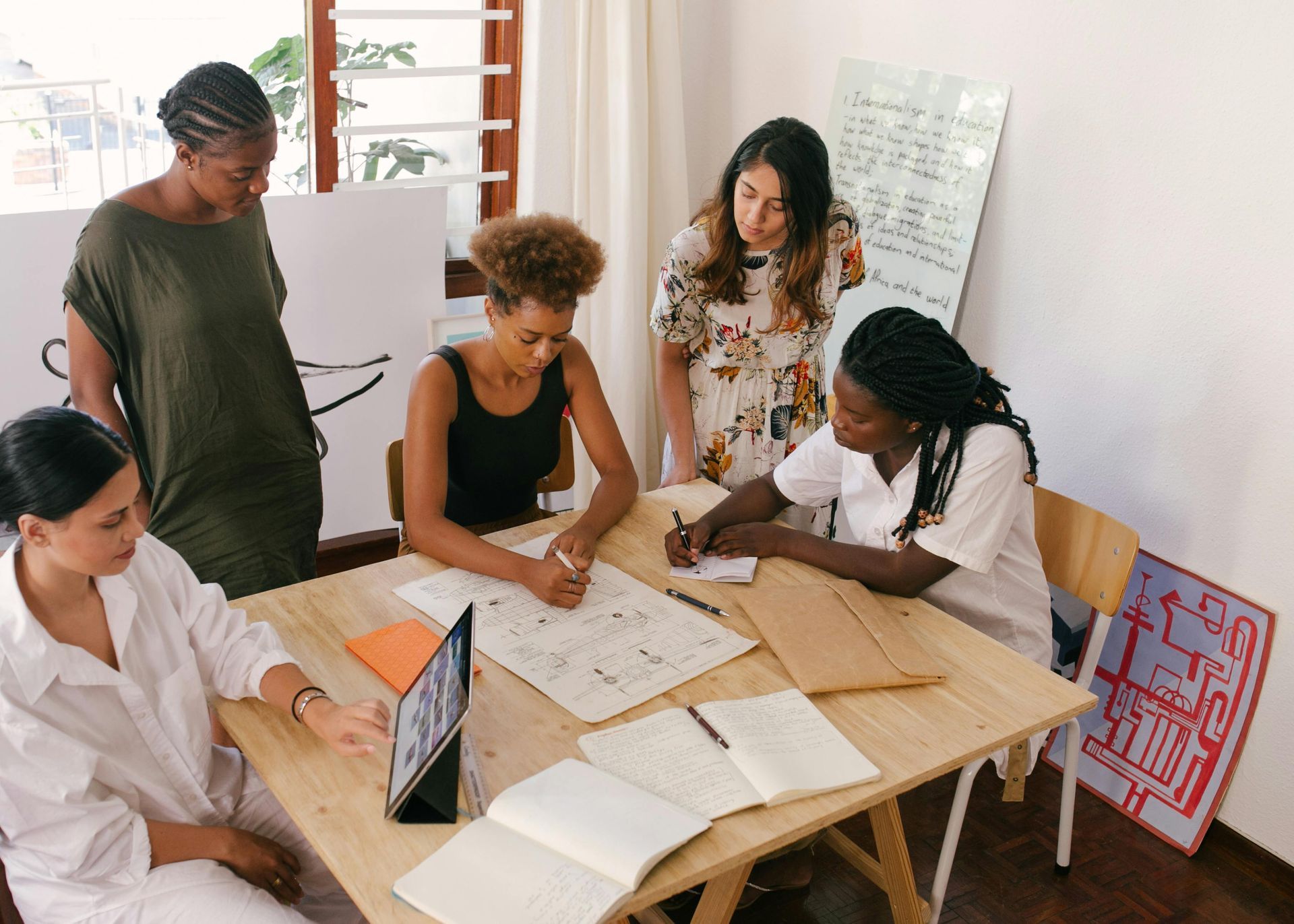 Five people, including one person writing, collaborate around a wooden table in a well-lit room.