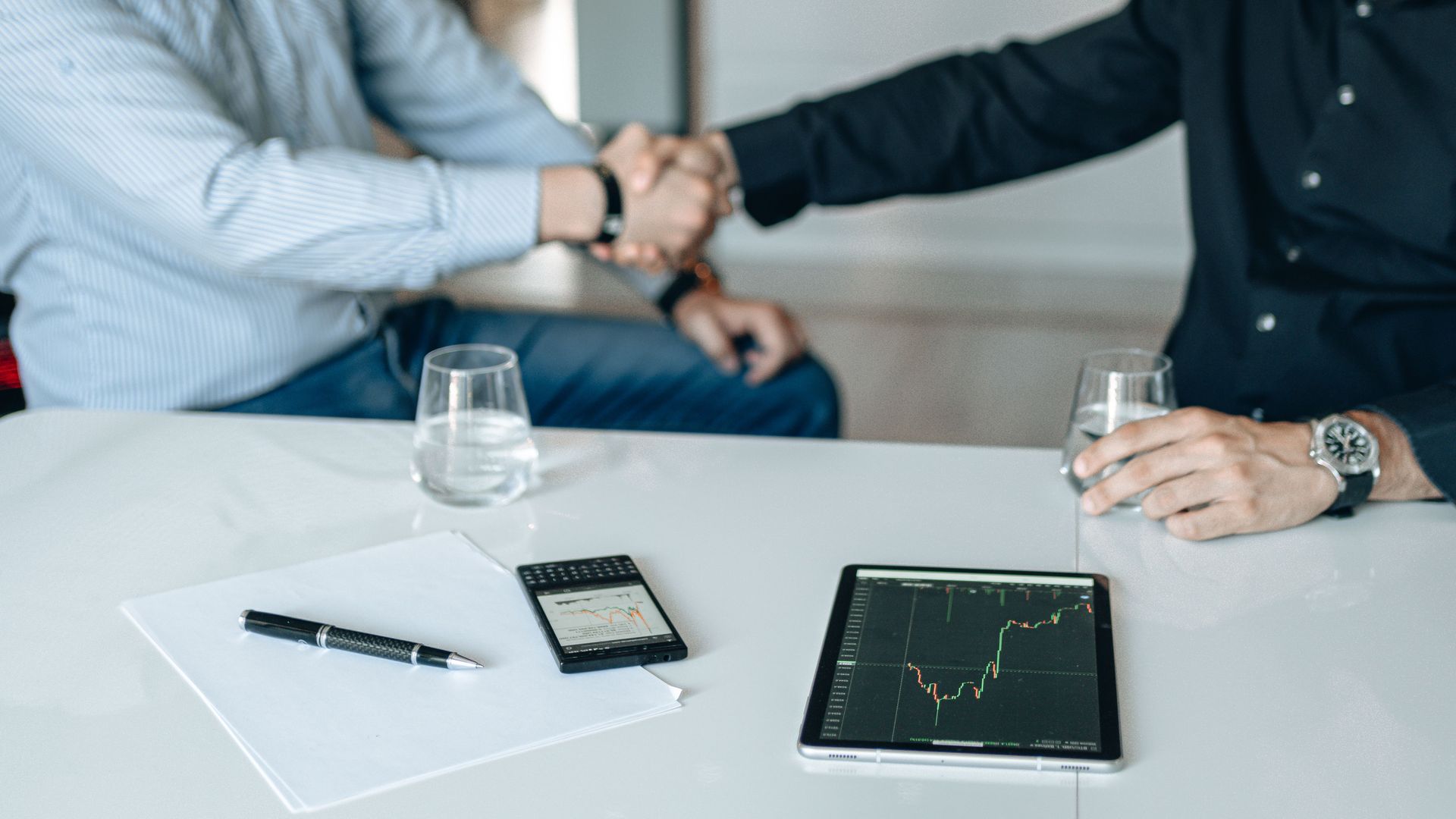 Two people shaking hands across a table with a tablet, pen, and glasses nearby.