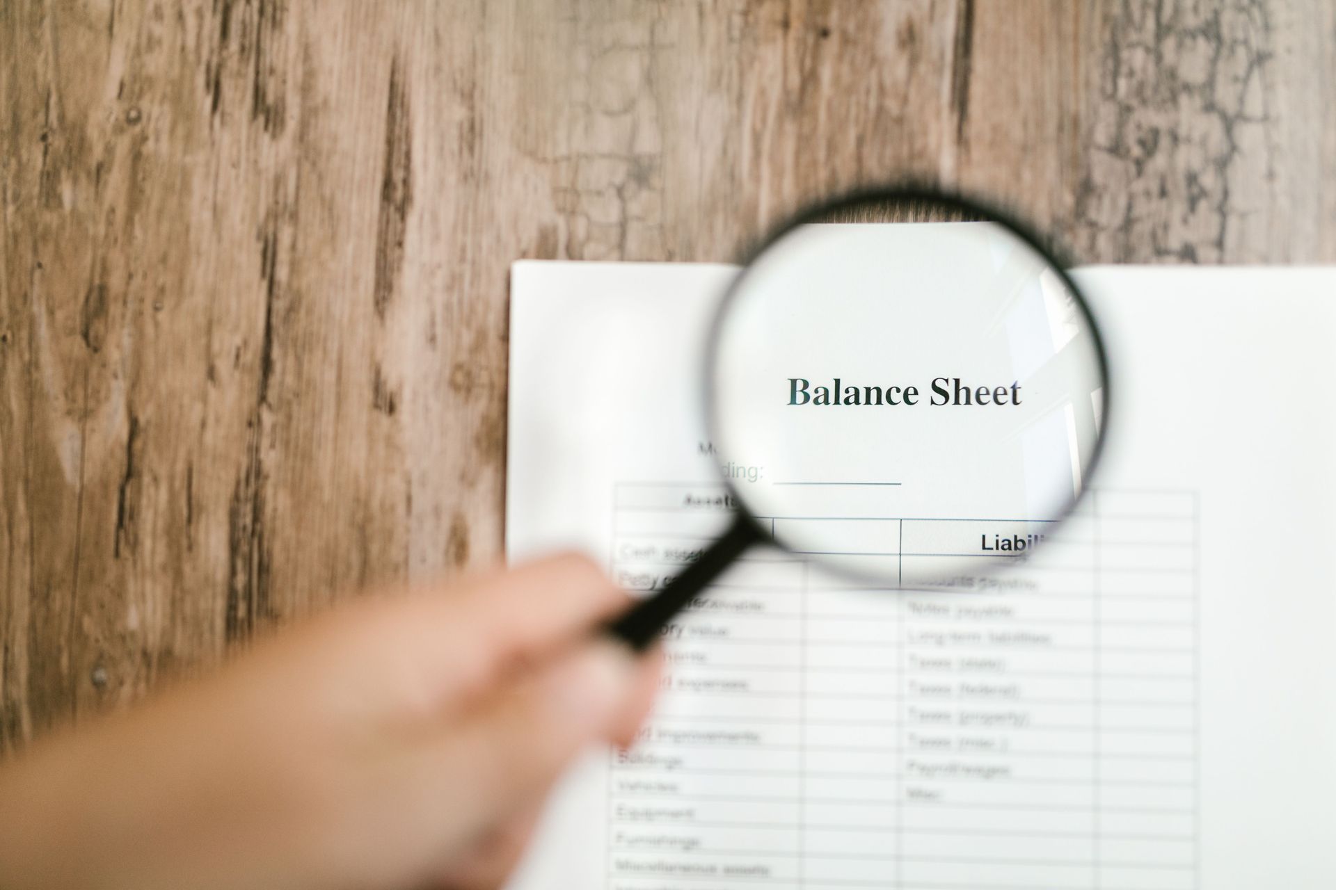Magnifying glass over a balance sheet on a wooden desk, with a blurred hand holding it