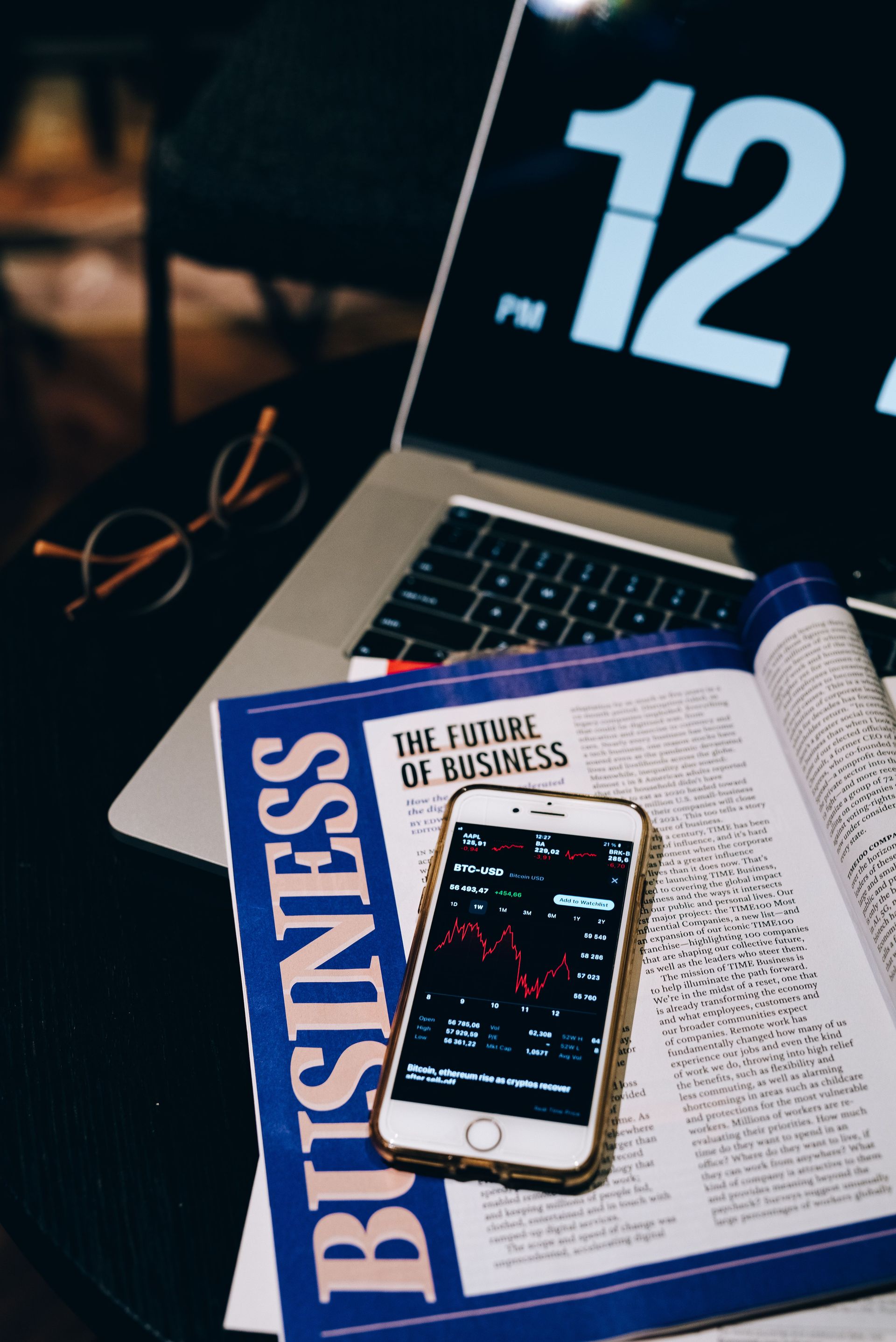 Business magazine and smartphone on a laptop keyboard with a clock display in the background