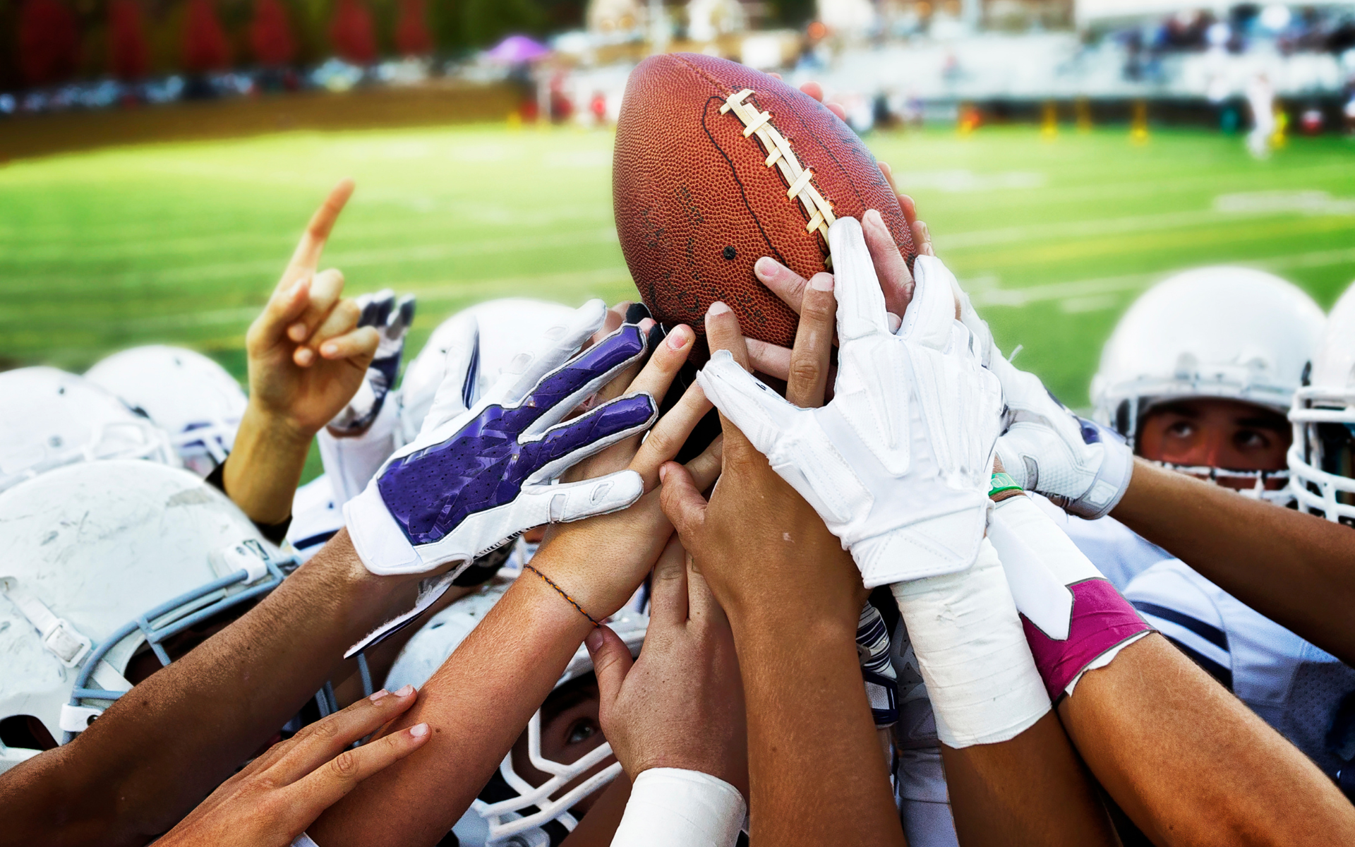 Football players in helmets and pads, hands stacked, holding the football on a green field.