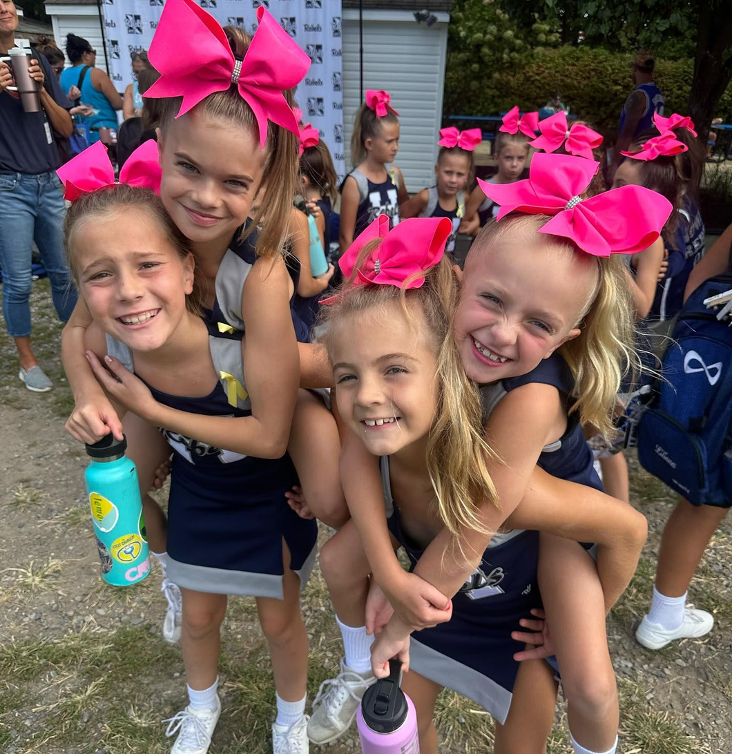 Group of young cheerleaders in navy and gray uniforms, smiling with pink bows.