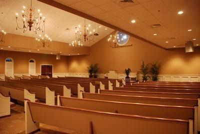 Interior of a church with rows of wooden pews facing a raised platform.  Tan walls, ornate chandeliers, and a circular window are visible.