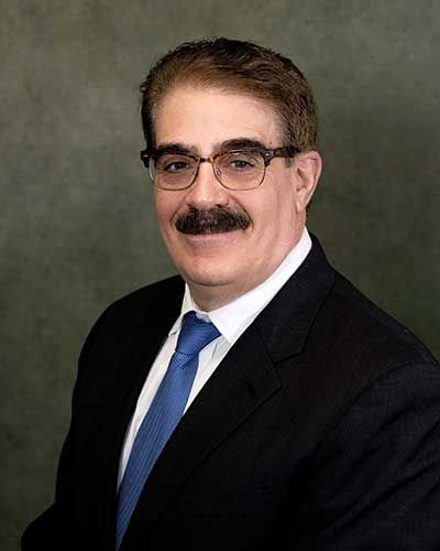 A professional headshot of a person with long, wavy brown hair, smiling, wearing a blue shirt under a dark blazer.