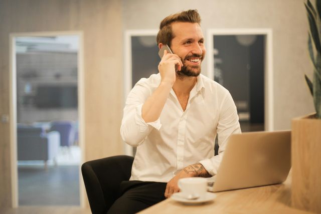 Man smiling while talking on a phone at a desk with a laptop and coffee in a modern office