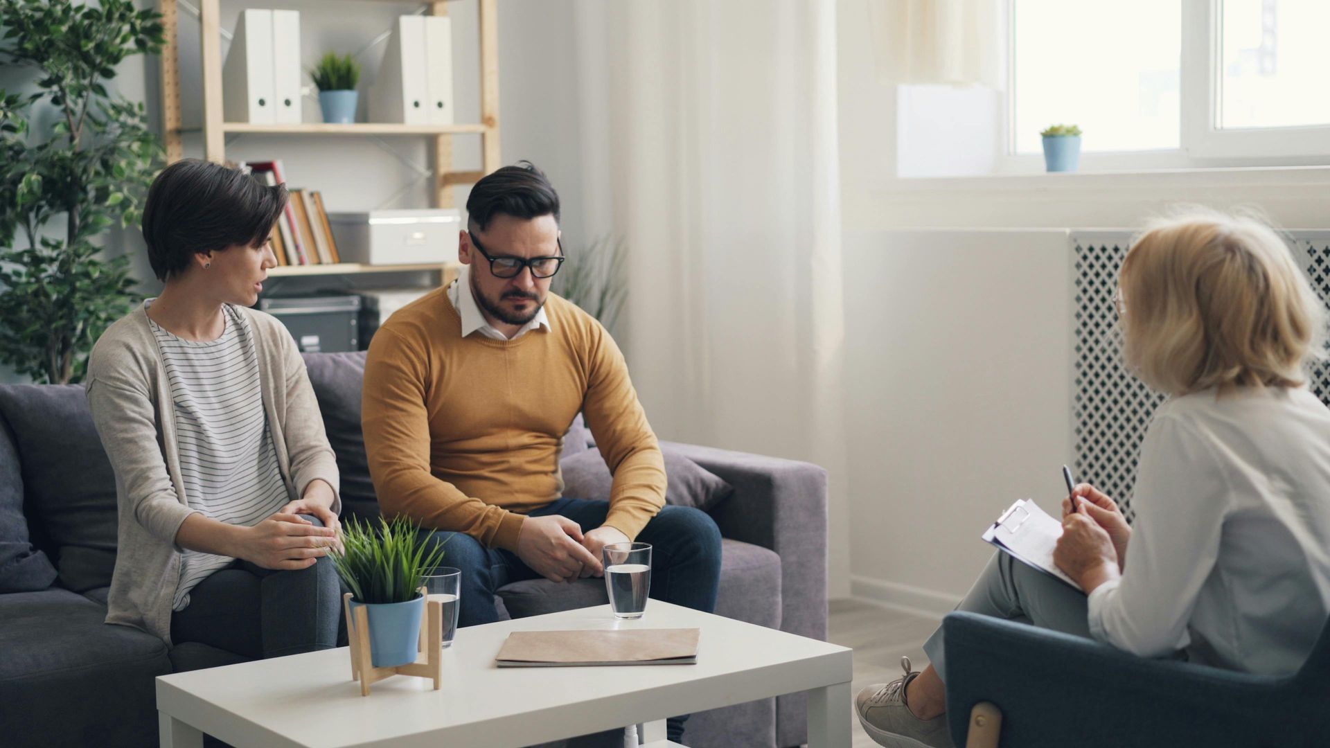 Elderly couple smiles while looking at a laptop on a couch. Woman has arm around man.