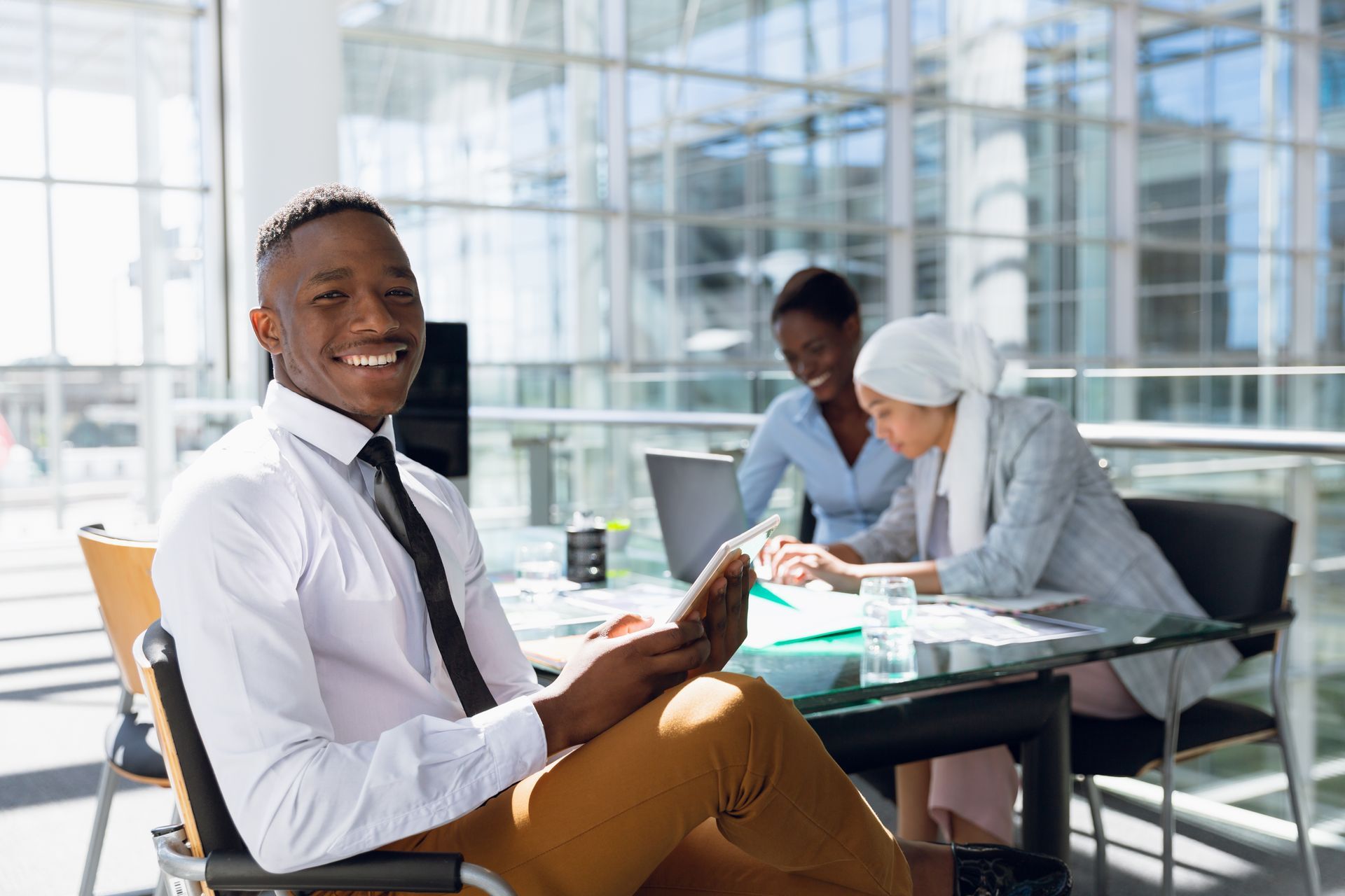 Man in office smiles, holding tablet. Two colleagues in background work at a table. Bright, modern office setting.
