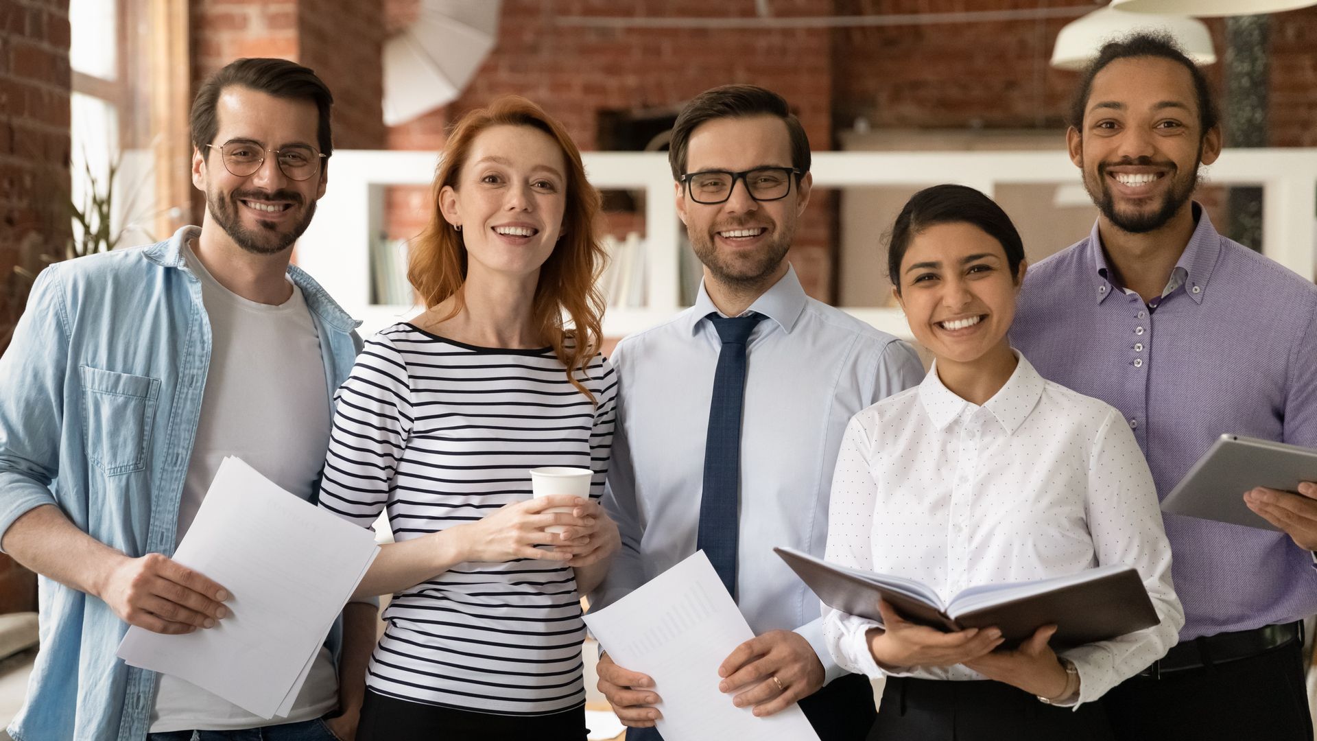 Office workers smiling, holding papers and a tablet. Brick wall background.